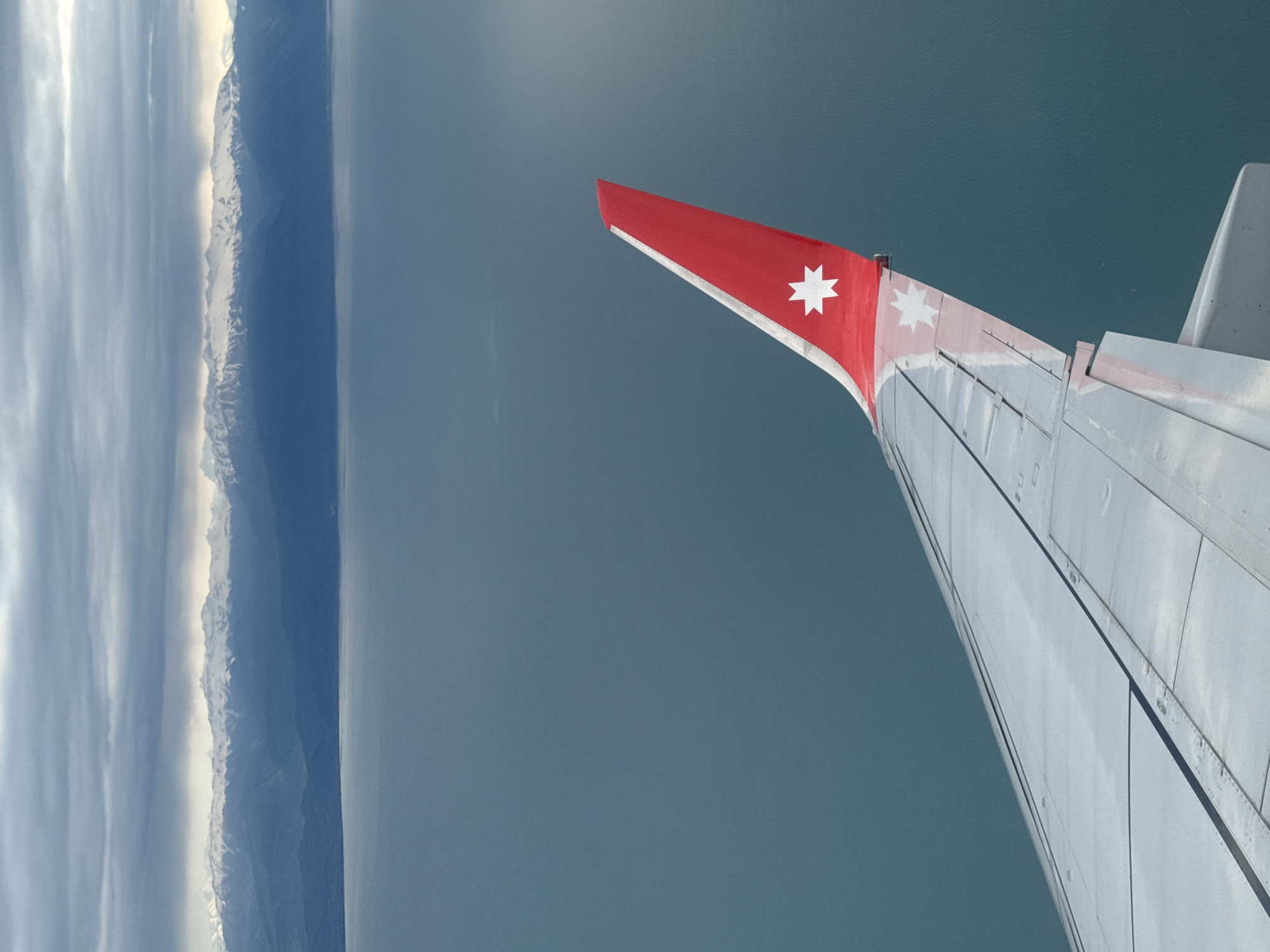 The image shows an airplane wing with a red tip and a white star, seen from inside the plane. Below the wing, there is a body of water. To the left of the water, there is a mountainous landscape with patches of snow on the peaks. The image is rotated 90 degrees clockwise. The location is not identifiable as Sochi, Russia.