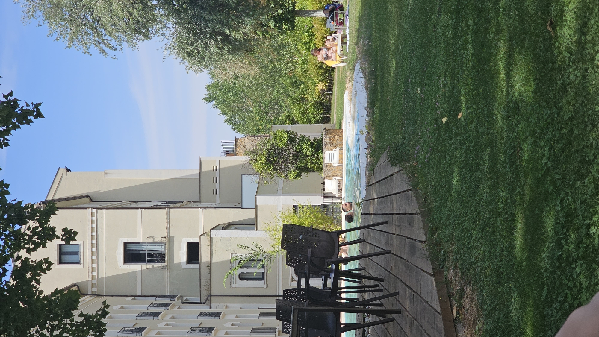 This media file captures a sunny day scene at what appears to be a resort or hotel in Alhama De Aragón, Spain. The primary focus is on an outdoor swimming pool area.
In the foreground, a wooden deck leads to the pool. Several black plastic chairs are arranged on the deck, some facing the pool and others angled away. The pool itself is visible, with tiled edges and clear blue water. Two people are partially submerged in the pool, with their heads and shoulders visible. One person, lying on their back, appears relaxed.
Beyond the pool, a well-maintained lawn stretches out, bordered by lush green trees. In the middle ground, several people are relaxing on lounge chairs on the grass. Some are wearing swimwear and appear to be sunbathing, indicating it's a warm day.
A large, multi-story building with a light-colored facade is situated to the left of the scene. The building has multiple windows, some with dark panes and others with metal gratings. Architectural details, such as decorative elements along the facade and balconies, are visible. Trees with dense foliage frame the building and extend towards the top of the frame.
The sky is a clear, bright blue, with a few wispy clouds. The sunlight suggests it is daytime, likely late morning or afternoon, given the shadows cast by the trees and the intensity of the light. The overall atmosphere is one of leisure and relaxation in a pleasant outdoor environment. No text is visible in the image.