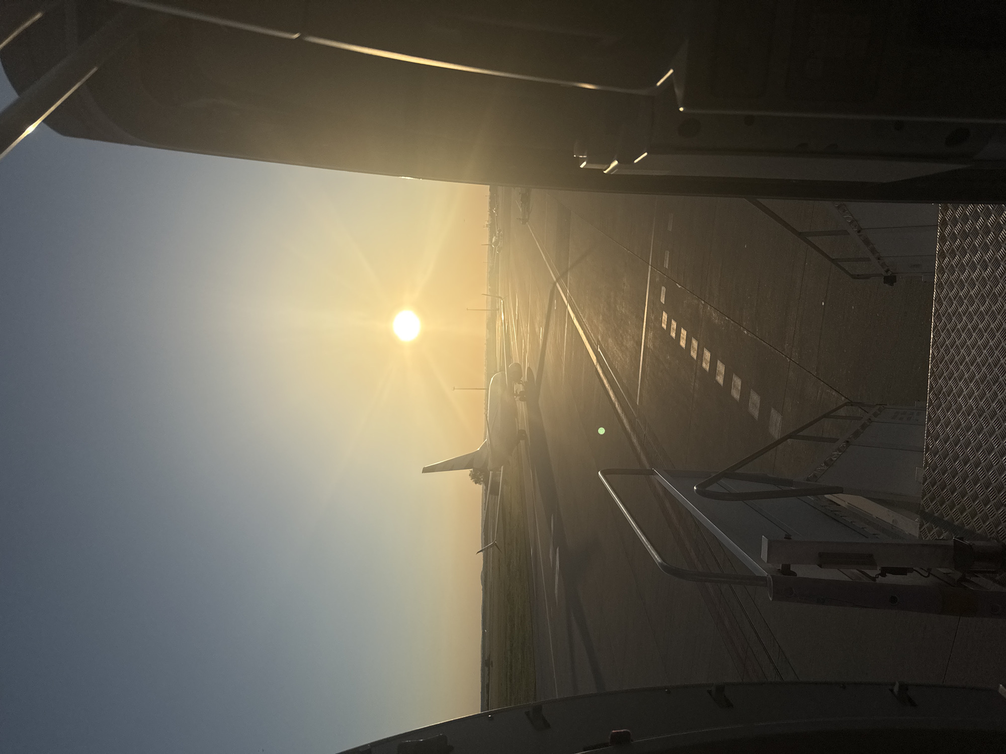 The image depicts an airport tarmac at sunrise, with the sun prominently displayed in the sky, casting a warm glow over the scene. The perspective appears to be from the top of an airstair, looking out onto the tarmac. Several airplanes are visible in the distance, parked on the runway. The foreground features the metal stairs and railings of the airstair. The sky is clear with a gradient of blue to yellow as it approaches the sun. The ground is asphalt, marked with various lines and squares, typical of an airport environment. There are no people or specific landmarks to identify the exact location in Prague, Czechia.