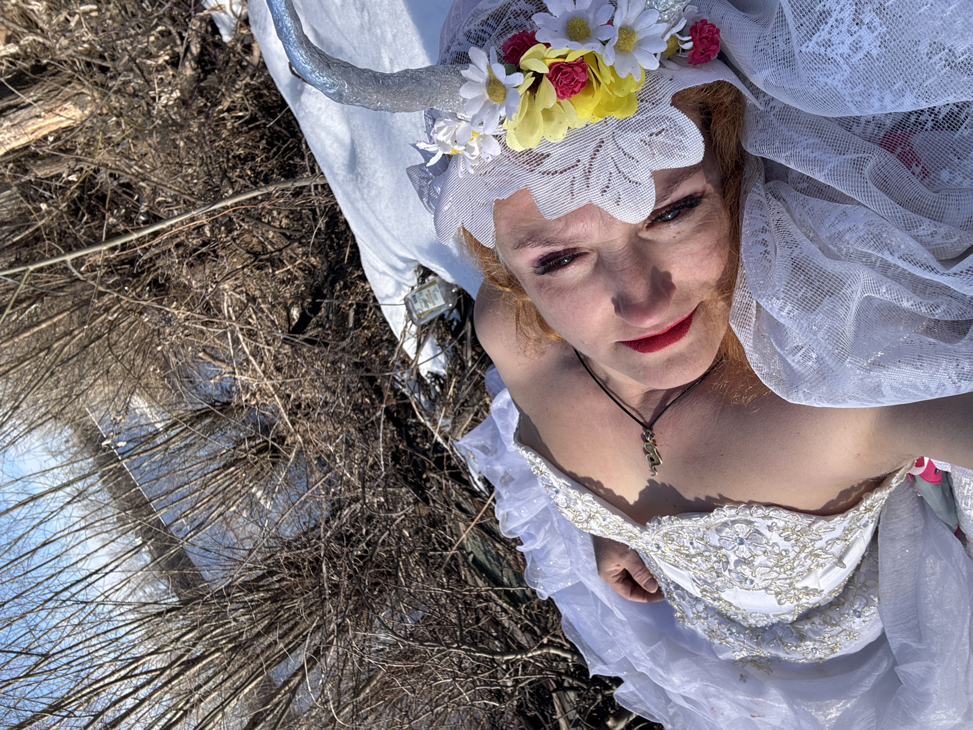 A woman in a white dress and headdress is positioned near a body of water surrounded by bare trees. The woman's headdress is adorned with flowers and silver horn-like attachments. The setting appears to be outdoors. Location: Chaikovsky, Russia.