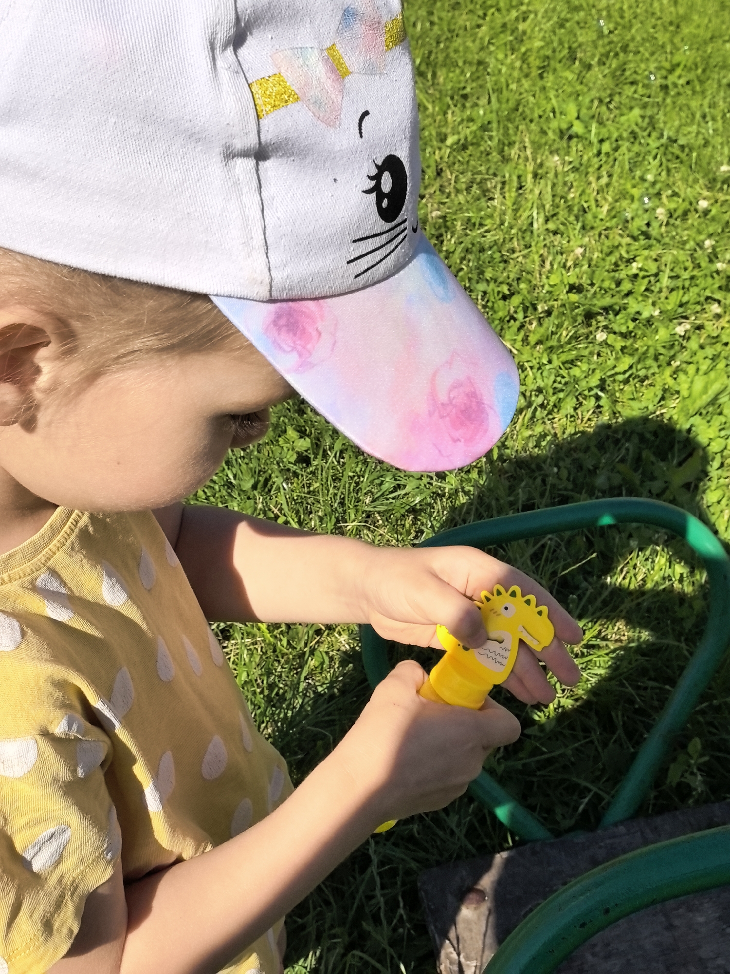 This media file captures a close-up view of a young child playing outdoors on a sunny day. The child, wearing a yellow polka-dot shirt and a white baseball cap with a cat design and pastel accents, is holding a yellow toy that appears to be a bubble blower shaped like a dinosaur. The child's hands are actively engaged with the toy.
The setting is a grassy area, indicative of a backyard or park, with sunlight casting shadows on the ground. A green hose is visible in the foreground, suggesting the presence of water or a watering system. The overall mood is one of playful innocence and enjoyment, typical of a child's outdoor activities. The time of day appears to be daytime, given the bright sunlight. No visible text is present in the media file.
