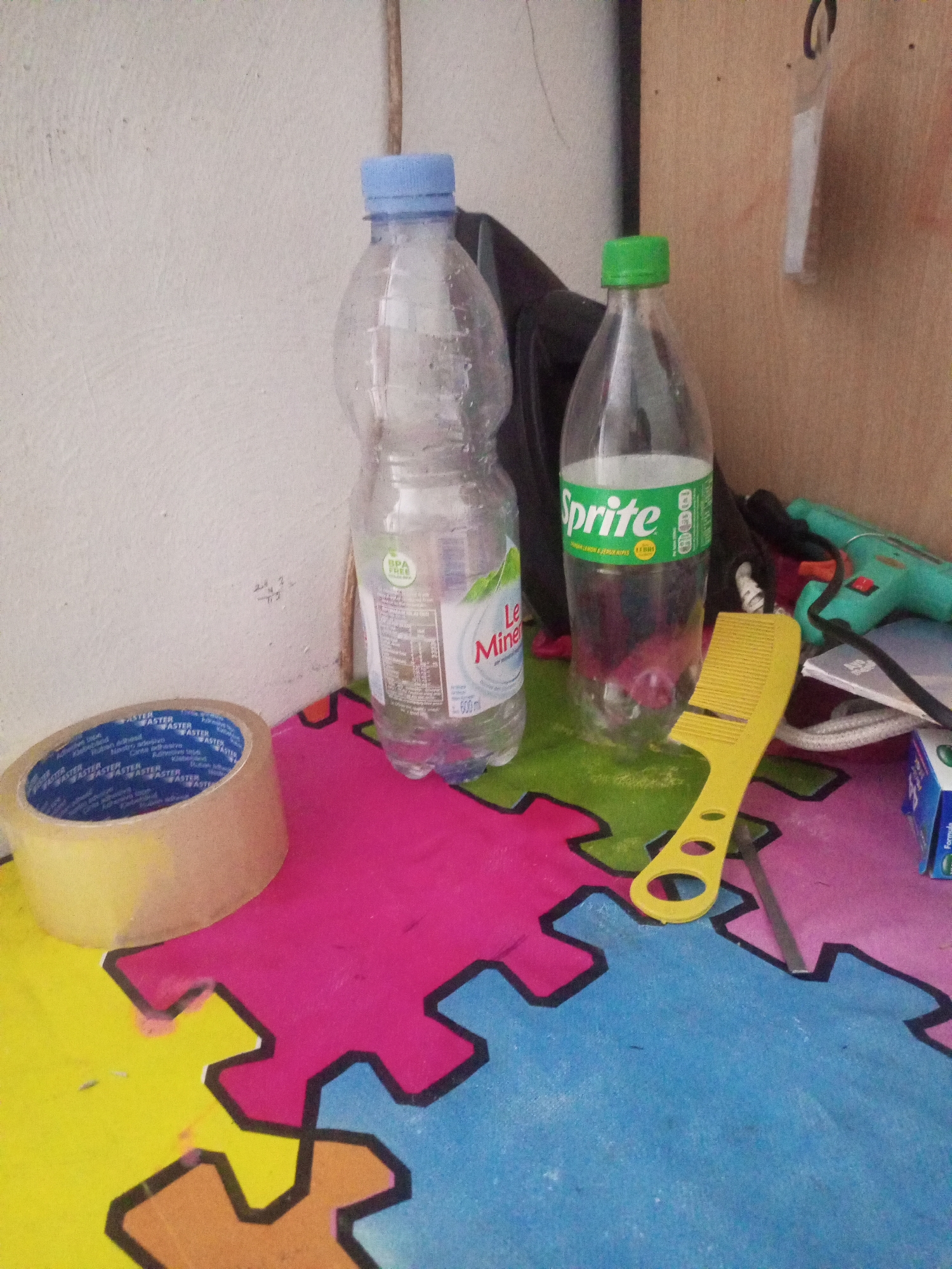 In an indoor setting with a white wall and a wooden surface, a collection of objects rests on a colorful interlocking mat. On the left, a roll of tan-colored tape with blue and white writing is positioned. Next to it, a clear plastic bottle with a blue cap contains a light-colored liquid. To the right of this bottle is a Sprite soda bottle with a green cap. In front of the Sprite bottle, a bright yellow plastic comb lies horizontally. Further to the right, a teal-colored electronic device, possibly a toy gun, is visible. A partially opened blue and white box is also present on the right side of the frame. The background includes a textured white wall and a wooden panel.