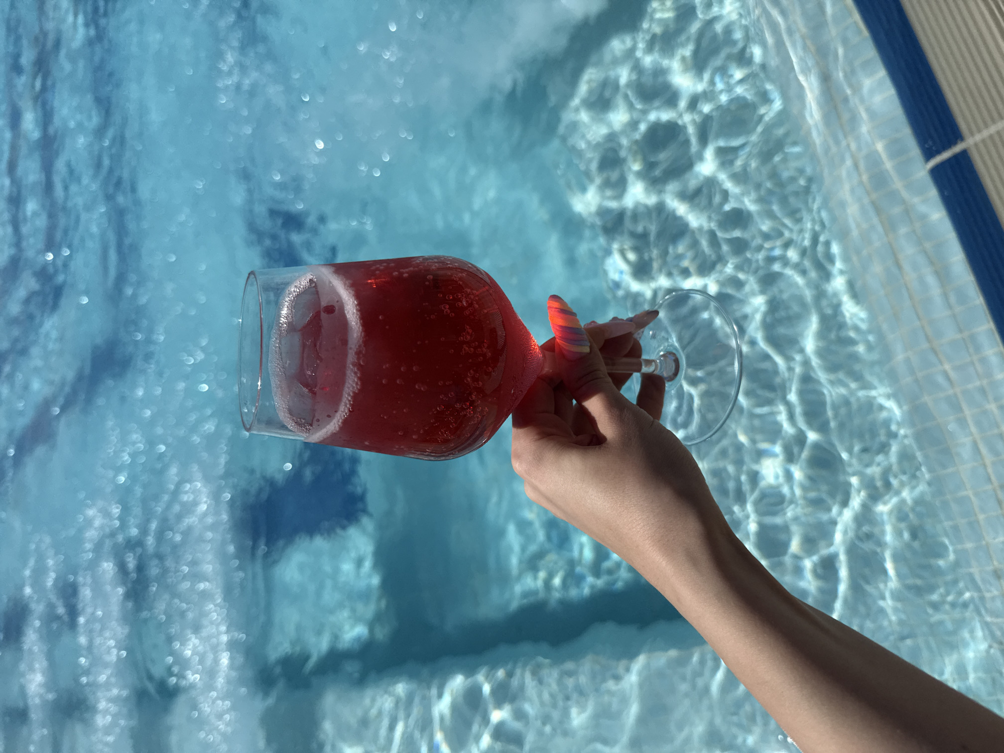 A fair-skinned left arm and hand are visible in the lower-left and central portion of the image, holding a clear, stemless glass containing a vibrant red, carbonated beverage. Small bubbles are visible within the liquid, with a thin layer of white foam at the top of the drink. The person's fingernails are long, manicured, and feature a multicolored, possibly rainbow-striped, design on the tips. The hand is positioned over a bright blue swimming pool, which occupies the majority of the background. The pool water exhibits strong light reflections and rippling patterns, indicative of direct sunlight. The pool's interior features light blue square tiles, with a distinct darker blue tiled line marking the edge of the pool in the lower-right corner. The water appears transparent and clear.