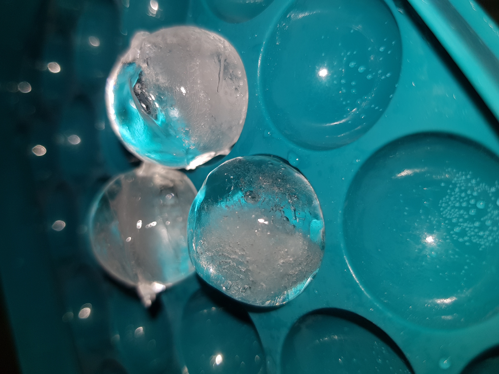 The image shows a turquoise silicone ice ball tray with three spherical ice balls. Some of the ice ball tray molds are filled with water. The ice balls appear to be partially melted with a layer of frosted ice on the outside. Background lighting creates bokeh effect.