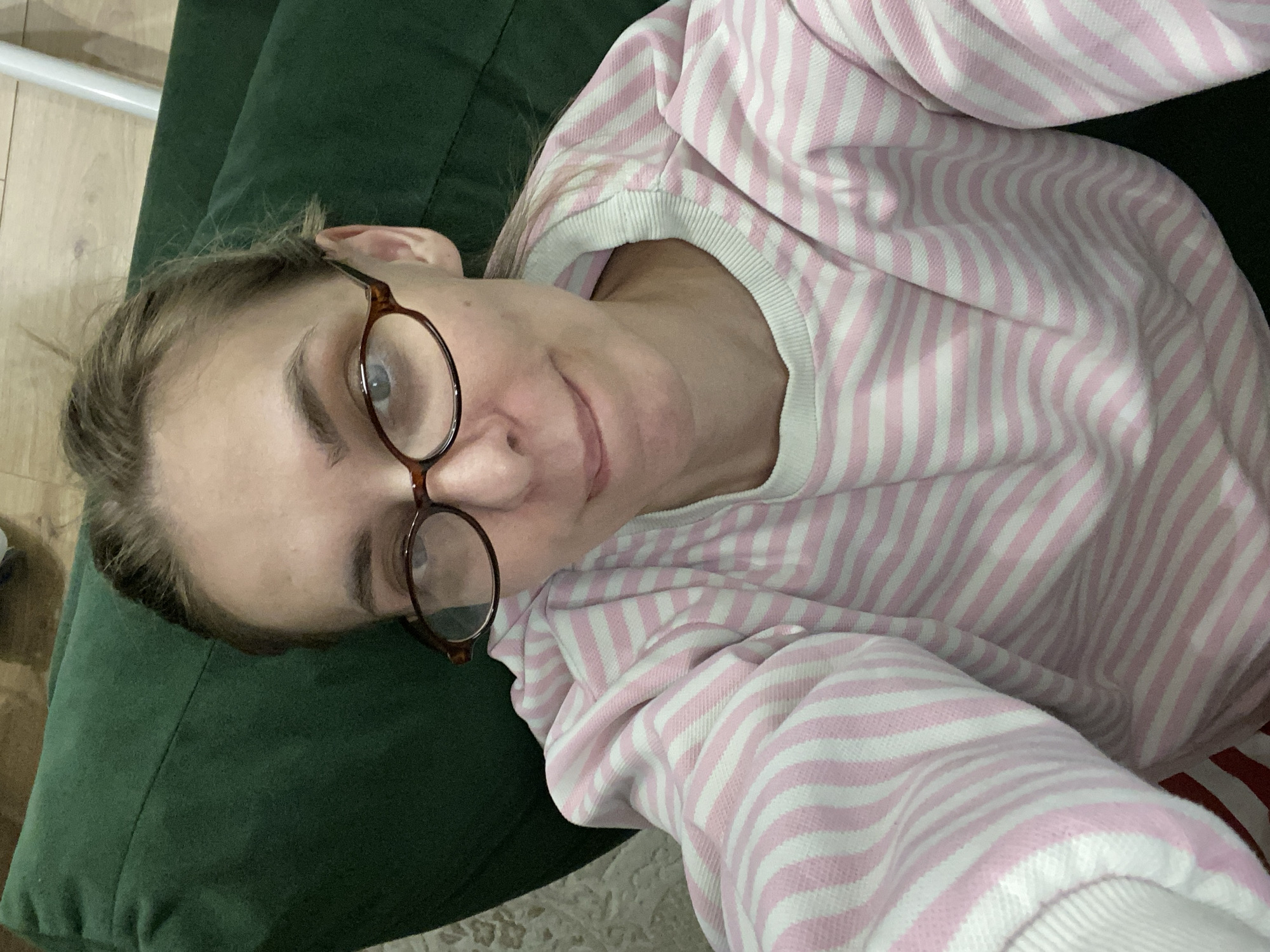 A single female subject, appearing to be in her late 20s to early 40s, is depicted in a high-angle selfie taken indoors, likely within a private residence in Chaikovsky, Russia. She has light-colored hair, light eyes, and is wearing brown, oval-framed eyeglasses. Her expression is a subtle, closed-lip smile. She wears a long-sleeved, crew-neck top with horizontal pink and white stripes. The subject is positioned against a dark green upholstered surface. Visible in the background are a light wooden floor on the right and a light-colored patterned rug on the left. A faint white vertical object is partially visible in the upper right background.