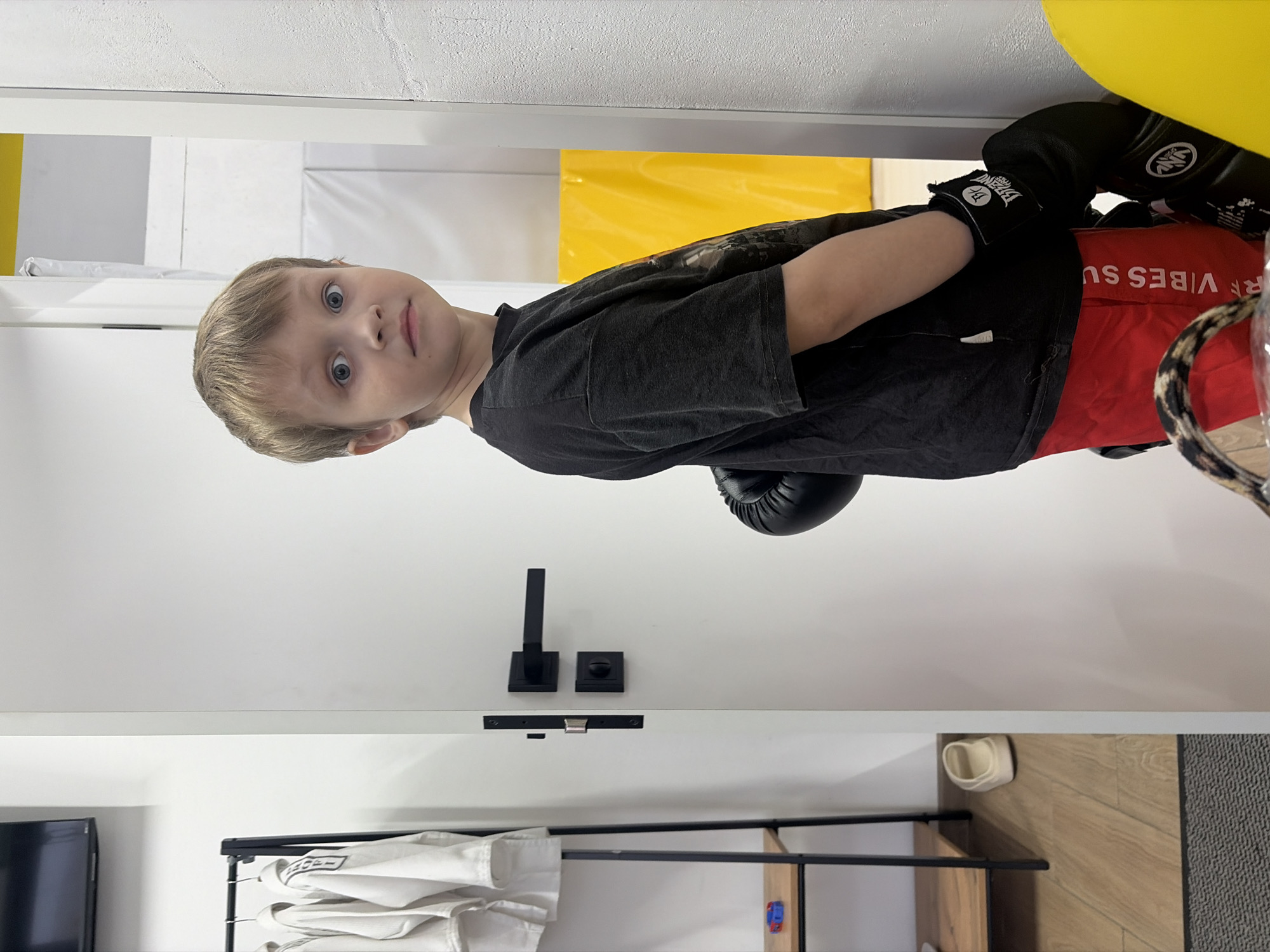 A young boy with blonde hair and blue eyes wearing boxing gloves and a dark t-shirt with red shorts is posing for a photo inside a building. The room is bright with white walls, and there is a clothing rack with white martial arts uniforms hanging on it. The location is difficult to determine specifically within St. Petersburg, Russia, as the image does not provide enough contextual clues such as landmarks or specific architectural details.