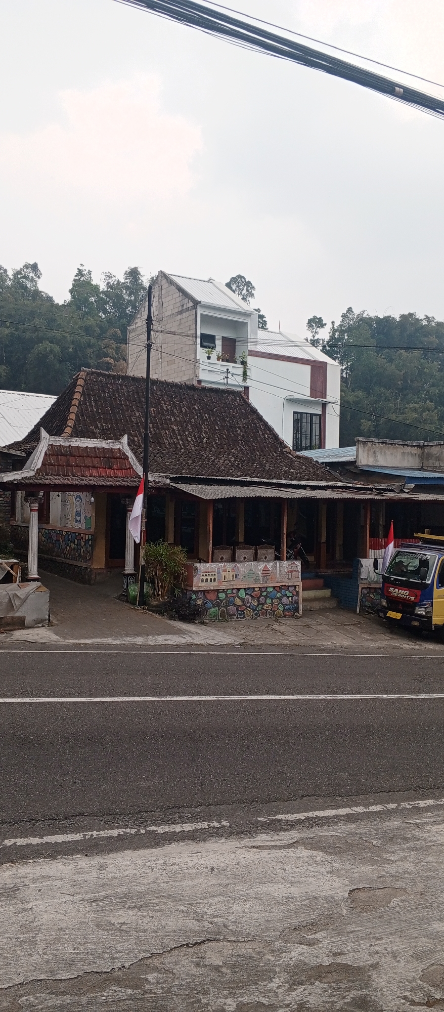 The image captures a street-level view of a building complex in Dadi, Indonesia, under an overcast sky. In the foreground, a paved road with white lane markings runs horizontally across the frame. Adjacent to the road is a concrete sidewalk.
The main structure in the center is a single-story building with a traditional tiled roof and an open-air seating area. The front facade of this building is decorated with colorful murals depicting abstract shapes, buildings, and figures. A flagpole with the Indonesian flag is planted in front of it.
Behind the single-story building, a multi-story modern building is visible. The lower portion is constructed of concrete blocks, and the upper sections are painted white with red accents and a distinctive angular roofline. Balconies with potted plants are present on one of the upper levels.
To the right of the main building, a yellow and blue truck is parked partially in the frame. The truck has lettering on its side, including "SANG PERINTIS". Various other structures with corrugated metal roofs are visible behind and to the sides of the main buildings, suggesting a densely built-up area.
The background consists of a dense forest with lush green trees, indicating a location that is likely not in the heart of a bustling urban center, but rather on the outskirts or in a more rural area of Dadi. The overall impression is one of a commercial or residential area situated near a natural landscape.