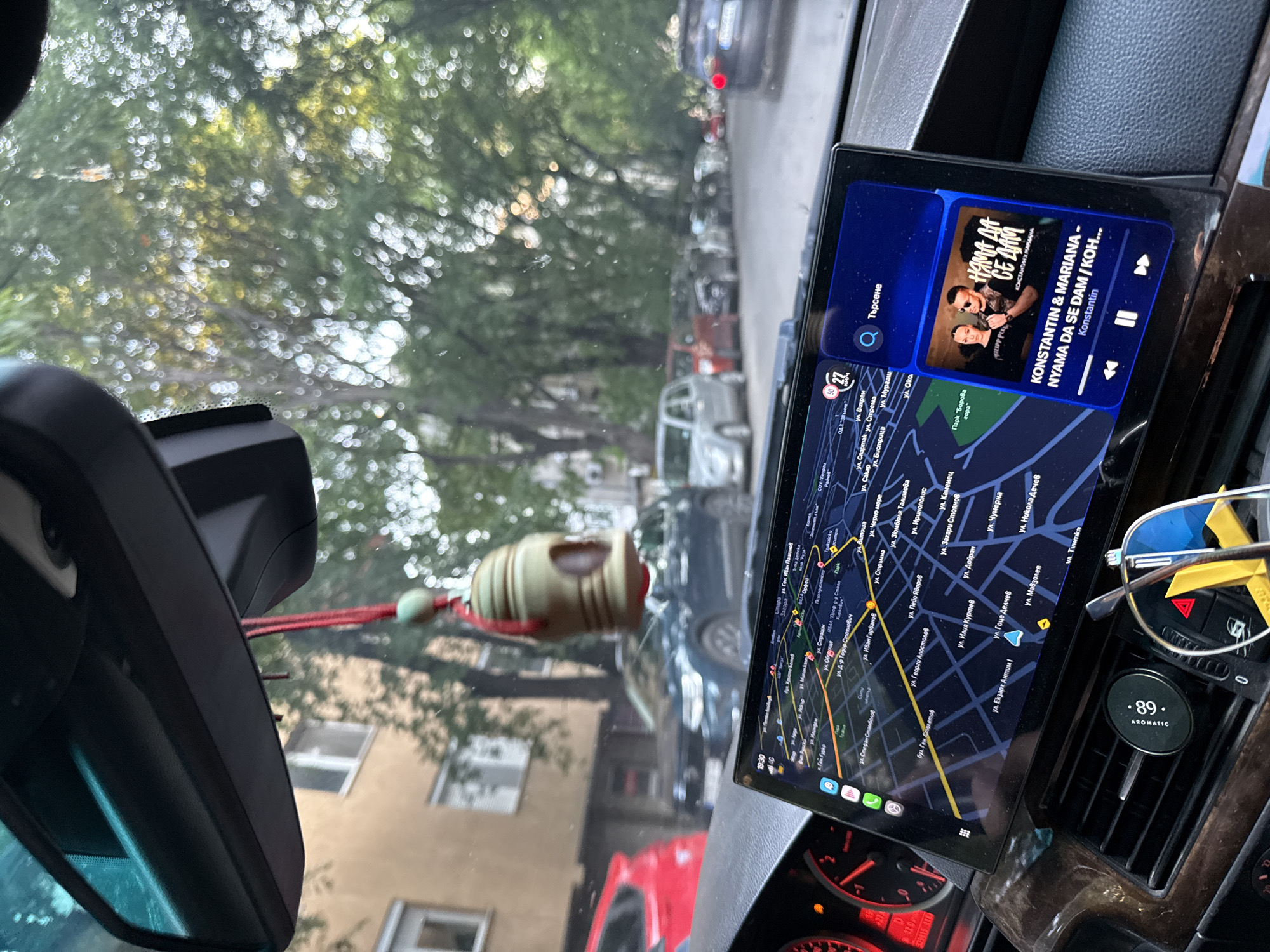 The image depicts the interior of a vehicle, viewed from a first-person perspective looking forward through the windshield onto an urban residential street in Stara Zagora, Bulgaria.
The lower portion of the image shows the car's dashboard, primarily dark with a wood-grain accent trim. On the far left, a portion of the instrument cluster is visible, illuminated with red lighting. A large, horizontally oriented infotainment screen is centrally mounted, displaying a navigation application on its left two-thirds and a music player interface on its right third.
The navigation map, rendered in shades of blue and green, displays numerous street names in Cyrillic, including "ул. Генерал Столетов" (Gen. Stoletov Street), "ул. Иван Гарванов" (Ivan Garvanov Street), "ул. Екзарх Антим I" (Exarch Antim I Street), "ул. Гоце Делчев" (Gotse Delchev Street), and "Парк Орфей" (Orpheus Park). The street names confirm the location as Stara Zagora. A highlighted route and a current location marker are visible on the map. The top left of the screen shows "19:30" (time) and a "4G" network indicator, alongside speed limit or speed indicators "50" and "27".
The music player section displays "KONSTANTIN & MARIANA - NYAMA DA SE DAM / KOH..." by Konstantin, accompanied by an album art image featuring two individuals. Playback controls (previous, play/pause, next) are present at the bottom of this section.
An "AROMATIC .89" branded circular device is attached to the right-side central air vent. Folded sunglasses with blue lenses and yellow frame accents rest nearby. A rearview mirror structure is visible at the top of the image, with a green bell-shaped charm hanging by a red string.
The exterior scene shows a narrow asphalt street with numerous parked and moving vehicles. On the immediate left foreground, the rear of a red hatchback is partially visible. Further down the street on the left, a dark blue SUV and a silver sedan are parked. Multiple additional vehicles are parked along both sides of the street, extending into the distance. A dark blue sedan with illuminated taillights is visible in the far-right background. Multi-story residential buildings with windows are visible behind the parked cars on the left. The street is heavily lined with mature trees featuring dense green foliage, forming a canopy overhead. Sunlight filters through the leaves, creating a soft, blurred background effect.