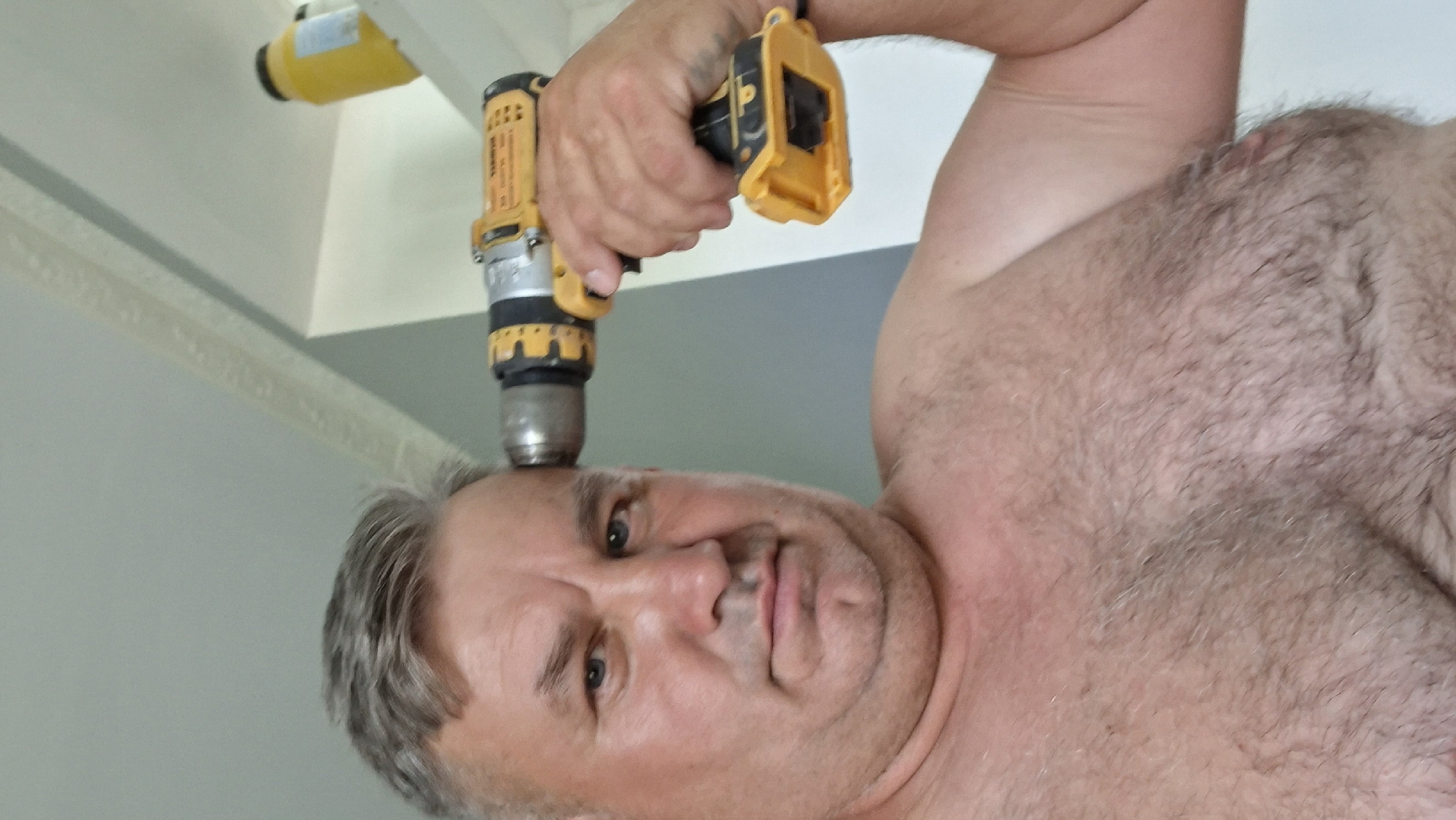 A shirtless, middle-aged man with graying hair holds a yellow and black power drill to his forehead. He is looking directly at the camera with a neutral expression. The background is a light-colored wall with some trim detail, and a yellow object is visible on a shelf in the upper right corner. The man's chest and shoulders are covered in hair. The image appears to be taken indoors, in what could be a home or workshop. The image is not identifiable as being in Voinova, Moldova.