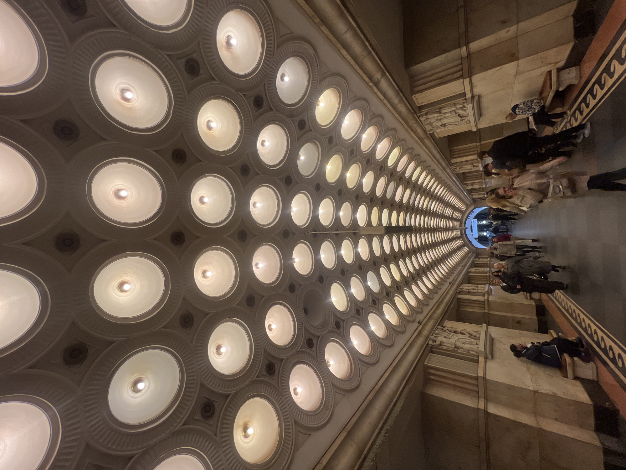 The image shows the interior of a metro station in an unknown city. The camera is looking upwards and down the length of the station. The ceiling is decorated with rows of round, recessed light fixtures, creating a tunnel of light stretching into the distance. The walls are constructed of stone or concrete, with architectural details such as cornices and friezes featuring relief sculptures. A number of people are visible standing or walking along the platform, with details of their clothing and posture suggesting a mix of locals and tourists. The lighting in the station is warm and diffuse, creating a soft glow throughout the space.