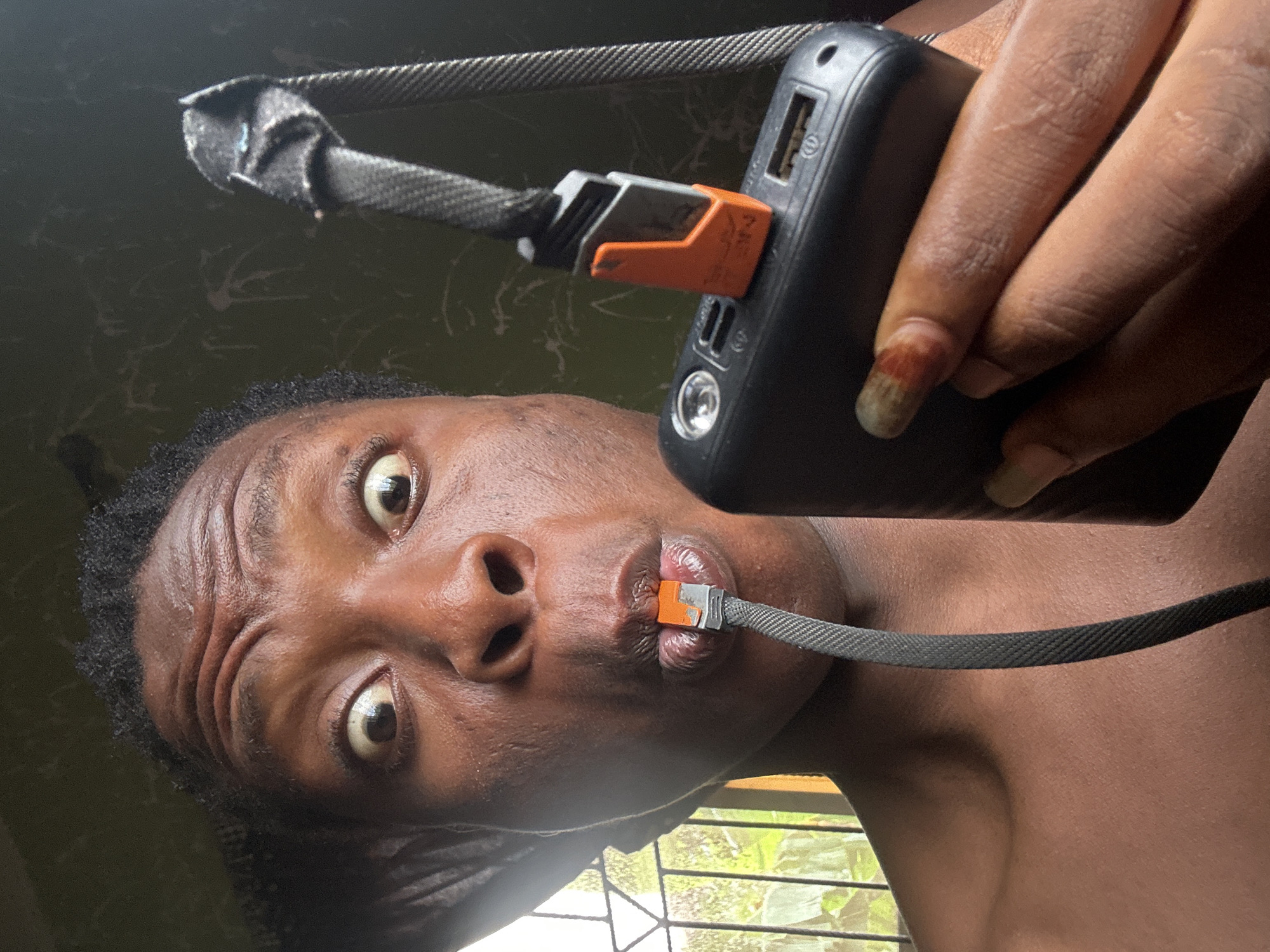 A person with dark skin is depicted in a close-up, tilted shot, looking upwards directly into the camera with wide eyes and a surprised expression. Their head is angled, with the right side lower than the left.
The person holds a black power bank in their right hand. A black braided charging cable, featuring orange and grey connectors, is plugged into one of the power bank's ports. The other end of the same cable, identical in connector design, is placed inside the person's mouth, with their lips slightly parted around it.
Visible details on the person include dark, curly hair, noticeable wrinkles on their forehead, and reflections in their eyes. The fingernail on their right index finger appears long and discolored, with shades of yellowish-brown and reddish hues.
The background is largely dark and indistinct, showing some subtle, light-colored smudges or patterns. Below the person's chin, a window is partially visible, revealing green foliage and horizontal bars or blinds outside. The lighting suggests an indoor setting with natural light entering from the window. There is no discernible text within the image.