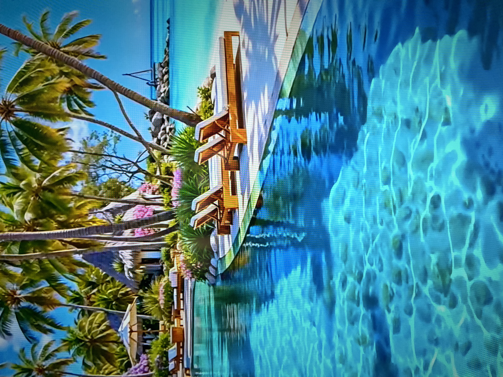 This is a digitally rendered image of a luxurious tropical resort scene, not a photograph of Kharkiv, Ukraine. The image depicts a pristine infinity pool with crystal-clear turquoise water in the foreground, reflecting the bright blue sky and palm trees.
Beyond the pool, a white sandy beach meets the ocean. Several wooden lounge chairs with white cushions are arranged on the beach, some facing the water. In the background, lush green palm trees tower over a cluster of thatched-roof bungalows, suggesting a secluded and tranquil getaway. Pink flowers add splashes of color to the landscape. To the right, rocks jut out into the ocean, and a small, triangular sail-like structure is visible on the water. The scene is bathed in bright sunlight, creating a vibrant and idyllic atmosphere.