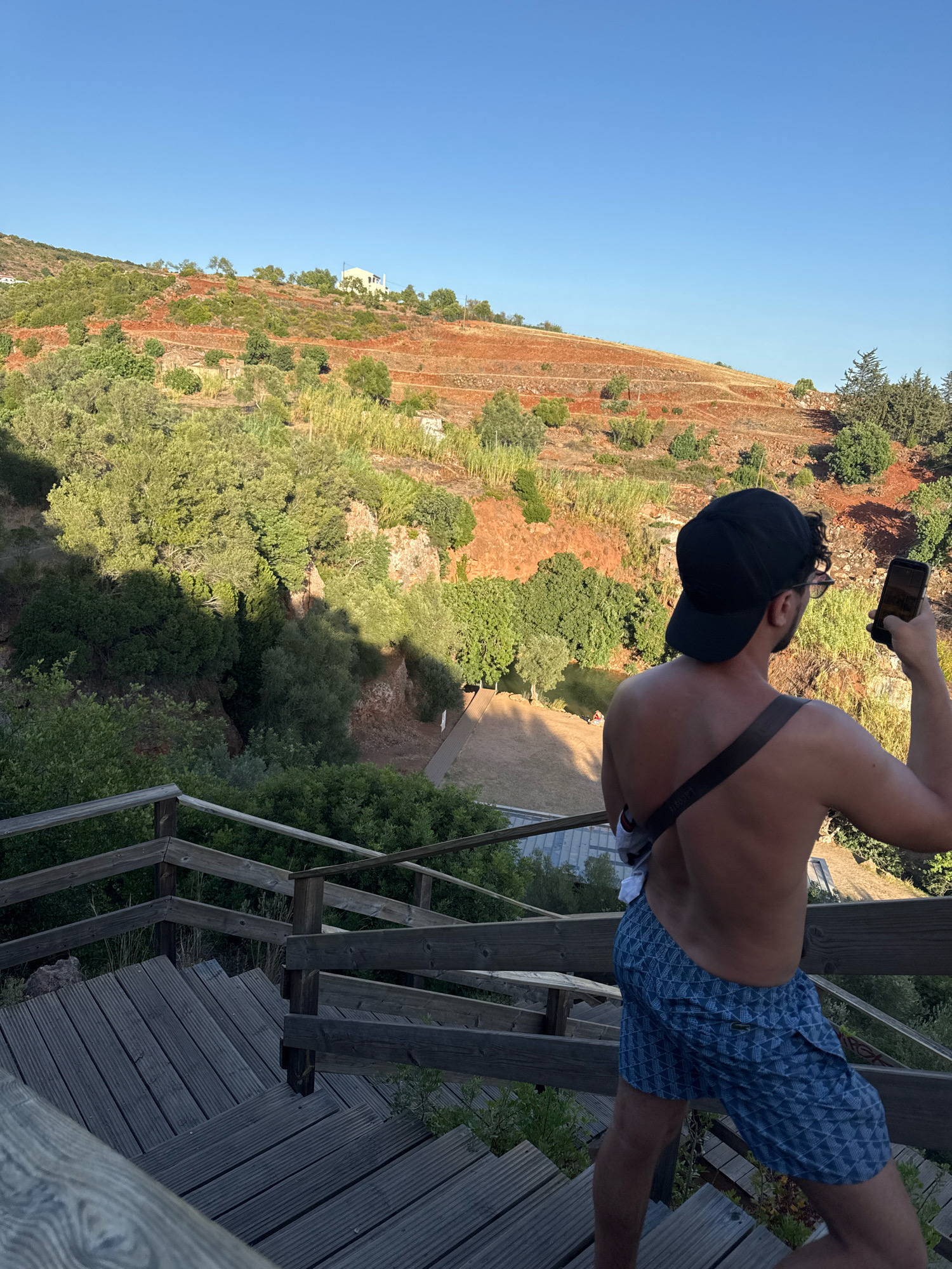 An adult male is pictured from the rear, standing on a wooden staircase. He is wearing a black baseball cap and blue patterned swim shorts. His back is bare, and a dark strap is slung across his shoulder. He holds a black smartphone in his right hand, appearing to take a photograph or video. The staircase is made of weathered wood, with railings on either side.
Beyond the staircase, a natural landscape unfolds. The ground is a rich reddish-brown earth, terraced in places, with patches of green vegetation, including trees and reeds. In the distance, a white building is visible on a hillside. A small body of water, possibly a river or pond, is situated in the middle ground, with a single person visible near its edge. The sky above is clear and blue. This scene is located in the Algarve region of Portugal, specifically within the municipality of Loulé, likely in an area known for its natural beauty and geological features, such as the Fonte da Benemola or nearby geologically interesting sites.