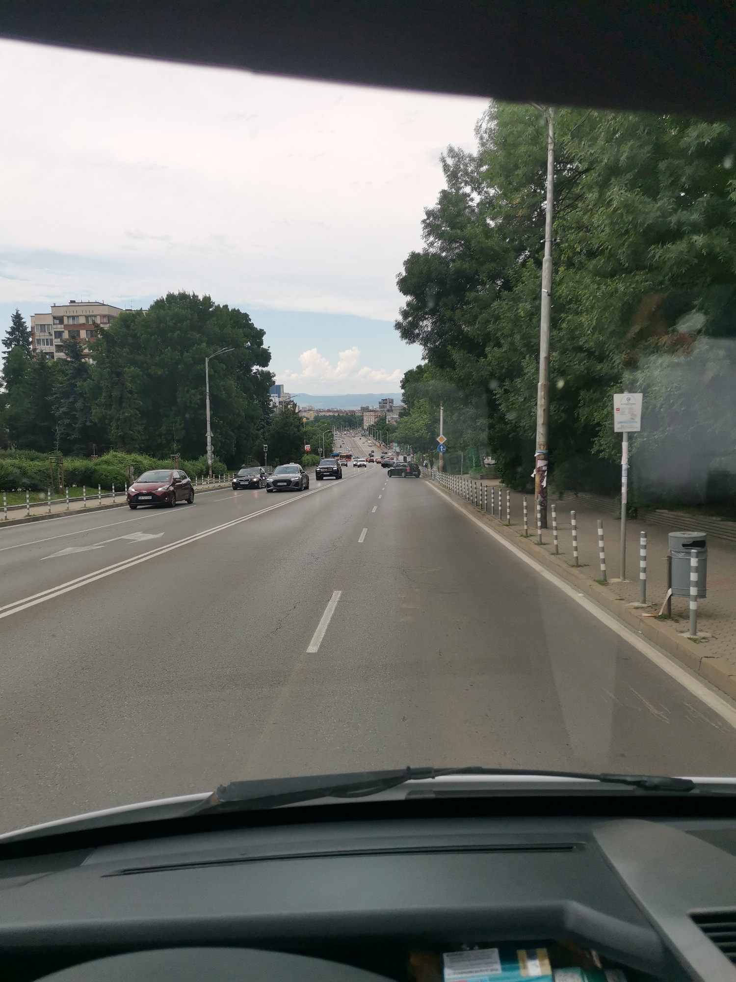 This image shows a view from inside a vehicle driving on a multi-lane road in Sofia, Bulgaria. The road has white lane markings and is bordered by a sidewalk with bollards and a trash bin on the right. Tall trees line the sidewalk, and beyond them, several residential buildings are visible. Other vehicles are on the road, including cars and what appear to be buses further in the distance. The sky is partly cloudy. The vehicle's dashboard and windshield wiper are visible in the foreground.