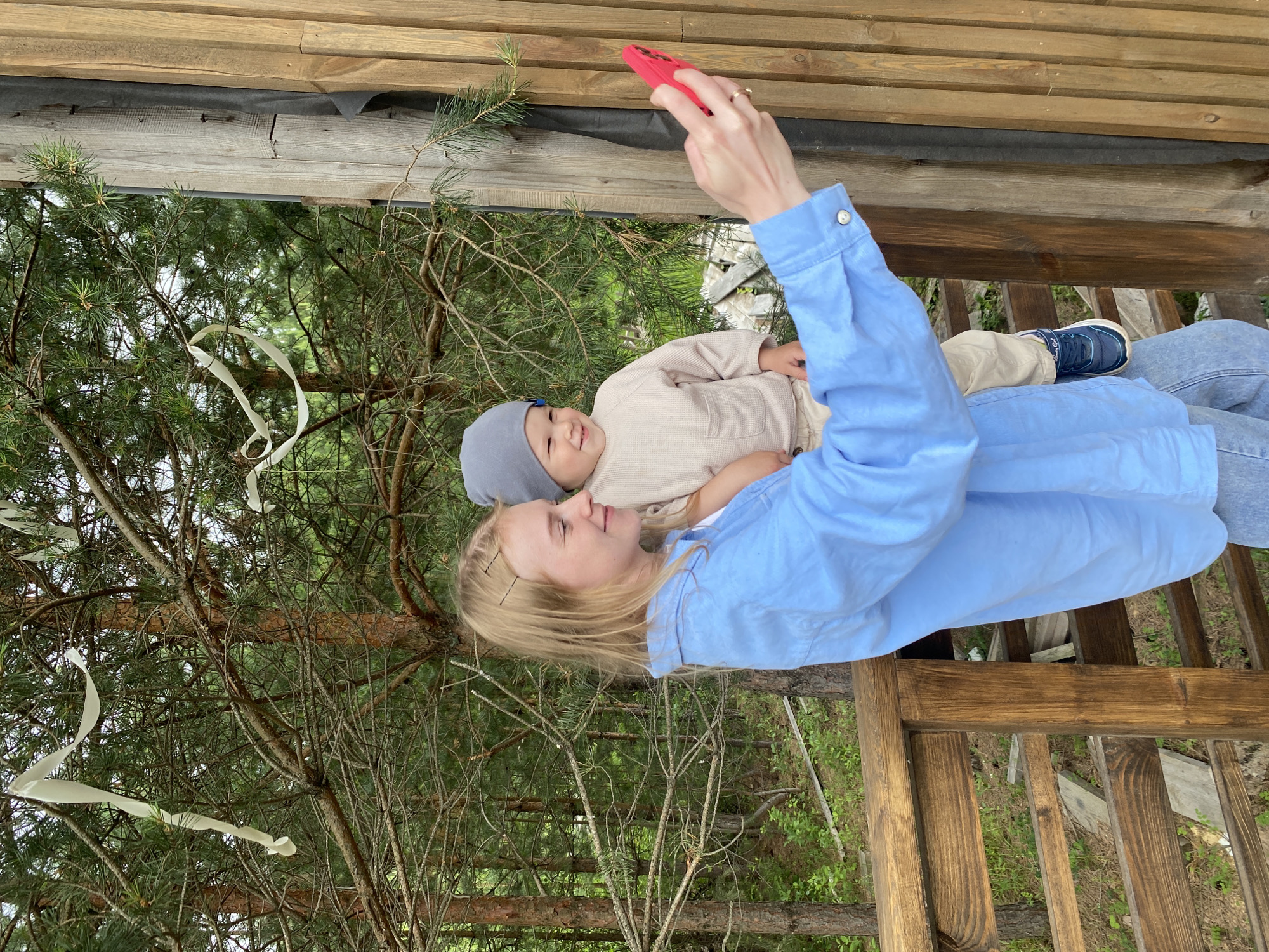 A young woman with blonde hair is taking a selfie with a baby in her arms on a wooden balcony. The woman is wearing a blue shirt and jeans, and the baby is wearing a beige onesie and a gray hat. They are in a wooded area, with pine trees visible in the background. There is a wooden railing in the foreground, and a wooden wall to the right of the woman. The image is taken from a medium shot, capturing the upper bodies of the woman and baby, and the surrounding environment. The lighting is natural and overcast. The location is identified as Fokino, Russia, based on the provided metadata.