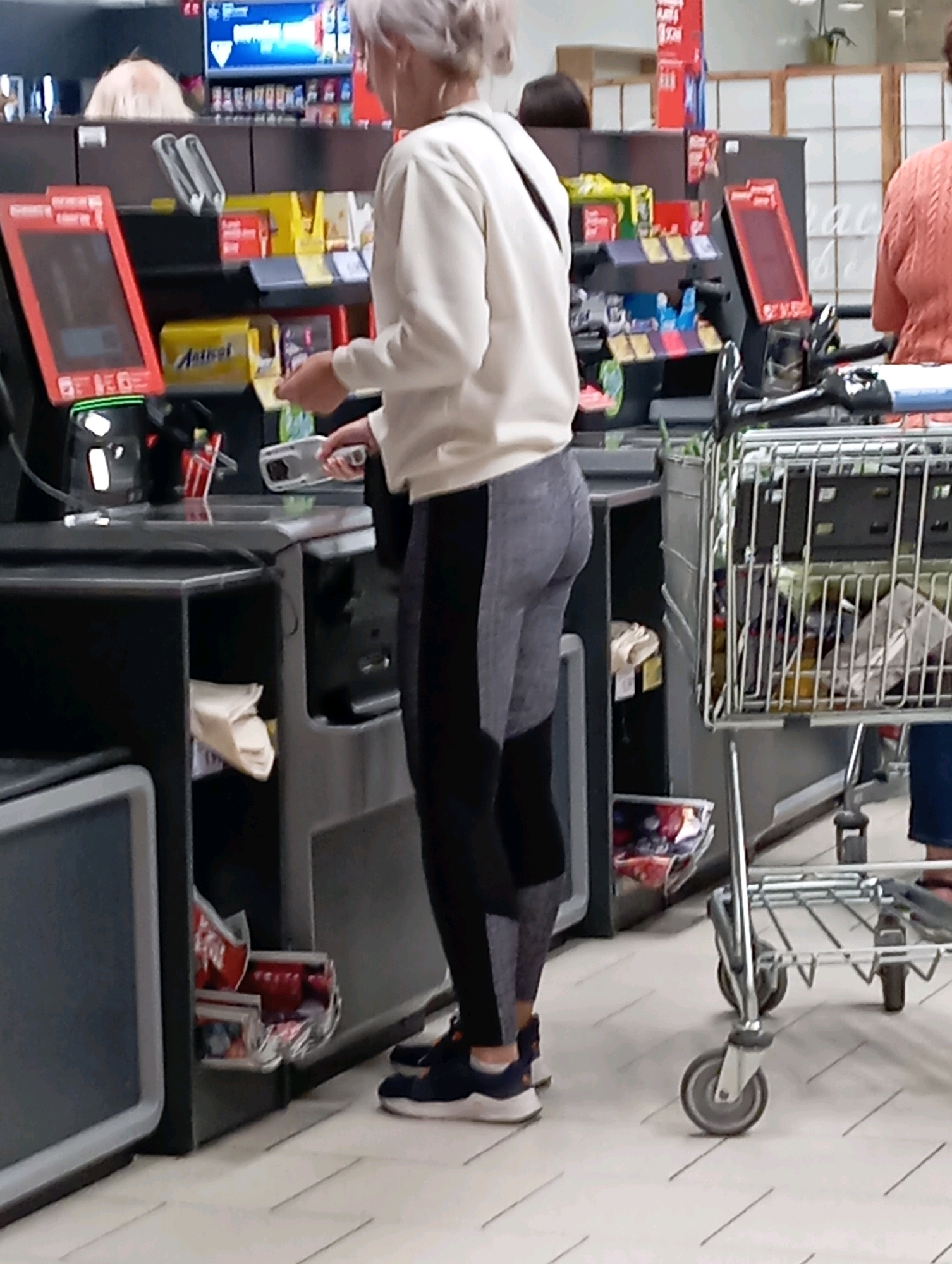 A person with blonde hair, wearing a white sweatshirt and grey and black leggings, stands at a self-checkout machine in a supermarket. The person is holding a scanner in their right hand and appears to be in the process of scanning an item. To the left of the person is a red self-checkout terminal. To the right, a shopping cart is visible, filled with groceries. The environment appears to be indoors, likely in a supermarket aisle, with shelving units stocked with products in the background. The flooring is tiled. The image does not provide any specific visual cues to determine the location in the city of Nitra, Slovakia.