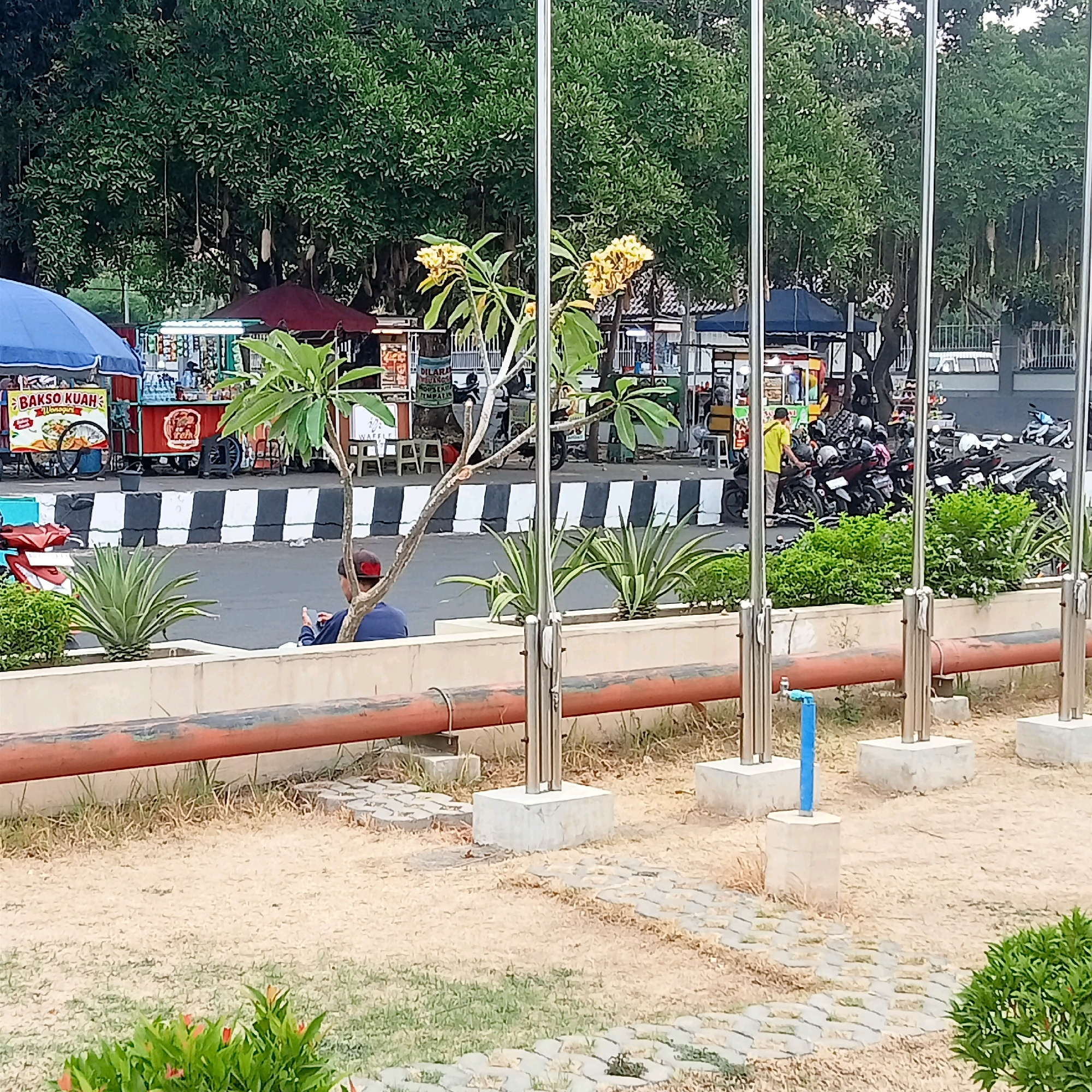 The image captures a street scene in the city of Karanganyar, Indonesia. In the foreground, a person is seated on a low wall, looking at their phone. Behind them, a street separates a row of food stalls from a grassy area with decorative plants and a large tree. The food stalls are open for business, with several motorcycles parked in front of them, suggesting a busy area. The overall environment appears to be an outdoor public space, possibly a park or pedestrian walkway adjacent to a road.