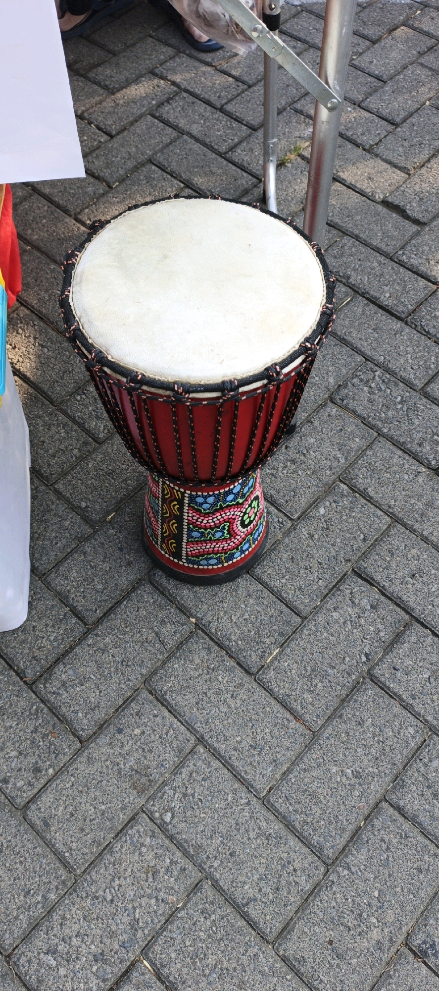 The image shows a close-up, high-angle view of a red djembe drum with a white drumhead, placed on a herringbone pattern of grey interlocking paving stones. The drum has black and red cord lacing around the top and a colorful, patterned base with designs in pink, blue, yellow, and black. To the left of the drum, a white plastic container is partially visible. In the background, a metallic support structure and a person's feet wearing dark shoes are visible. The lighting suggests it is daytime, with shadows cast by the drum and the support structure. The scene appears to be outdoors on a paved area. This location is identified as Sidoarjo, Indonesia.