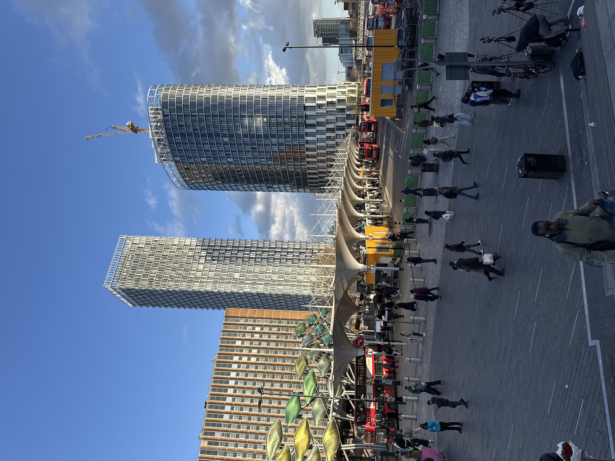 The image captures a bustling public square in London, United Kingdom, dominated by modern high-rise buildings. Two prominent glass-clad towers stand tall, with a construction crane visible atop one. Below, a covered walkway shelters a row of red double-decker buses and pedestrians. People are walking across the paved square, some carrying bags or engaging in conversation. Street furniture, including benches, trash receptacles, and what appears to be artistic metal structures, are scattered throughout the area. The sky is partly cloudy with blue patches.