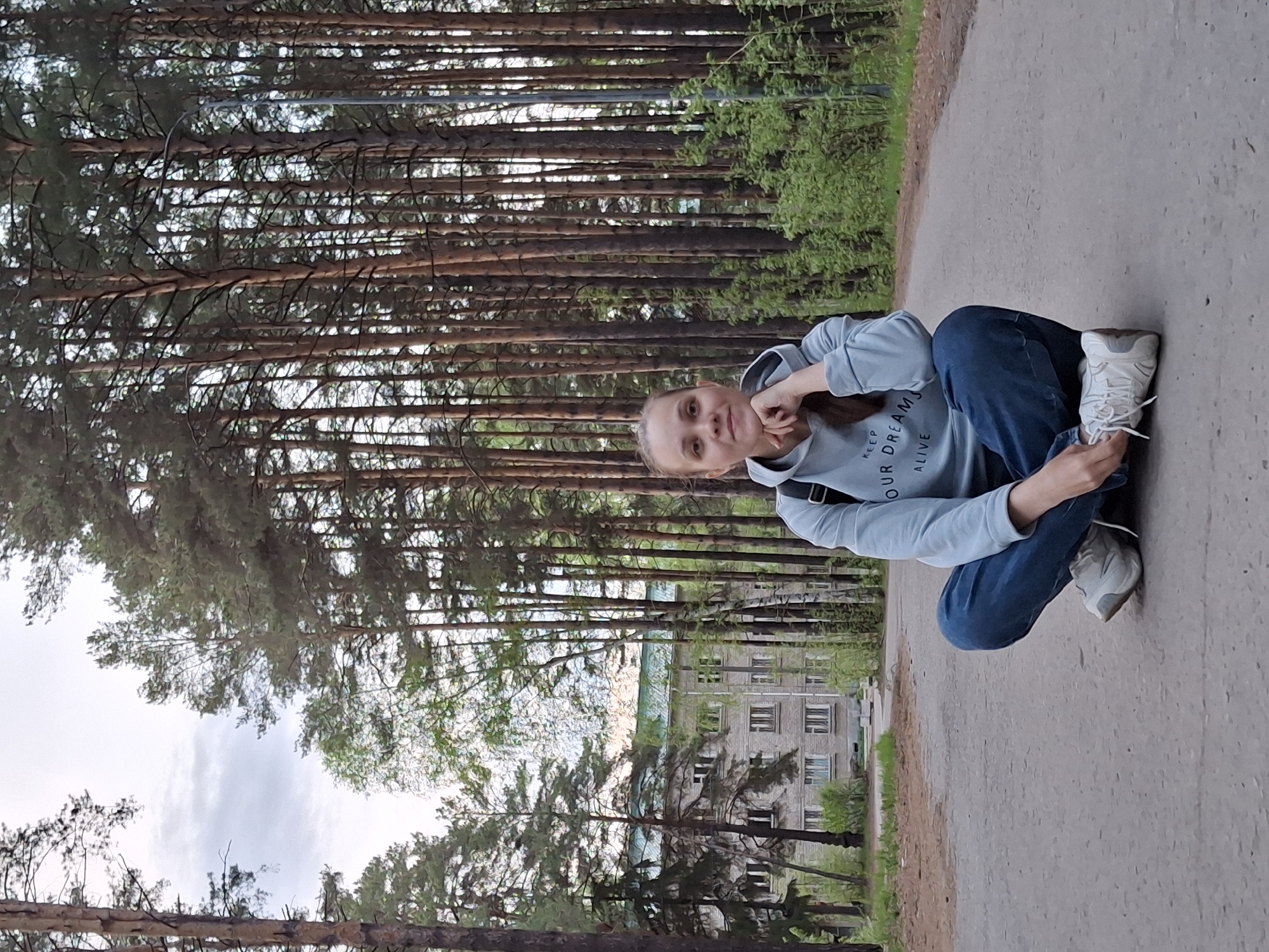 A woman is squatting on a paved surface, tying her shoelace. She is wearing blue jeans and a light blue hooded sweatshirt. She is facing forward, slightly turned to the left with her head cocked towards the camera. The background consists of tall pine trees, a building in the distance, and a grey sky. Based on the image content, this is most likely located on a path near a wooded area in the city of Chaikovsky, Russia.