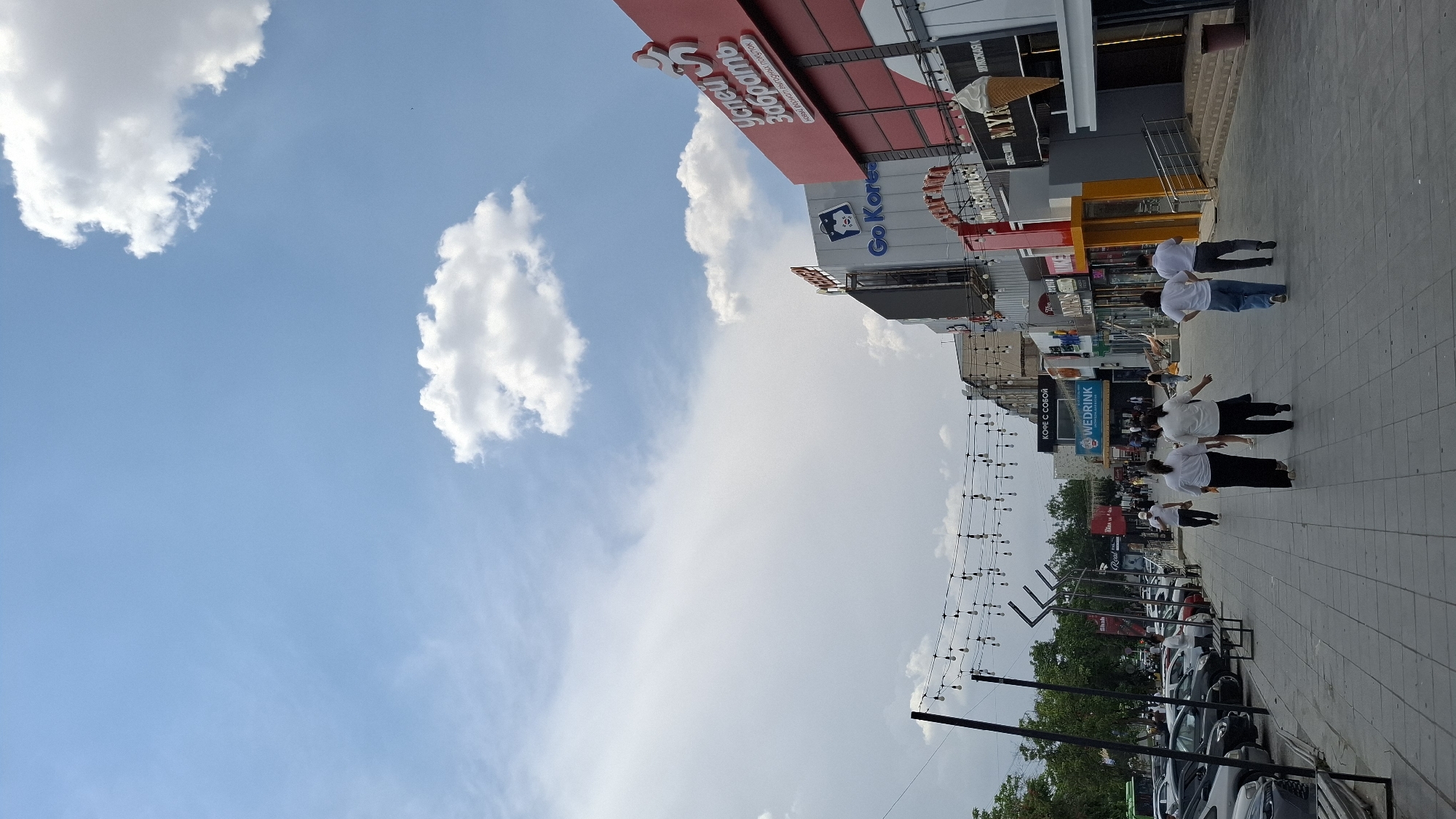 This is a horizontal, eye-level shot of a street scene in Shymkent, Kazakhstan, taken on a sunny day with scattered clouds. The image is oriented vertically, making it appear as though the viewer is looking down at the scene.
The foreground is a paved sidewalk lined with shops and businesses. Several people are walking on the sidewalk, most of them wearing white shirts and dark pants. One person in the distance is wearing a white shirt and blue jeans. Overhead, strings of lights are strung across the street.
Several businesses are visible with signs in Russian and English. One prominent sign reads "Успей Забрать" (Oospey Zabrat') which translates to "Hurry to Collect" or "Don't Miss Out". Another sign says "Go Korea", suggesting a business related to Korean products or services. "WEDRINK" is also visible on a sign, likely for a beverage establishment. An ice cream cone is depicted on top of one building.
Further down the street, parked cars are visible along the roadside. Trees and foliage can be seen beyond the buildings, suggesting an urban setting with some greenery. The bright sunlight and blue sky indicate it is daytime. The overall atmosphere appears to be that of a bustling commercial area.
