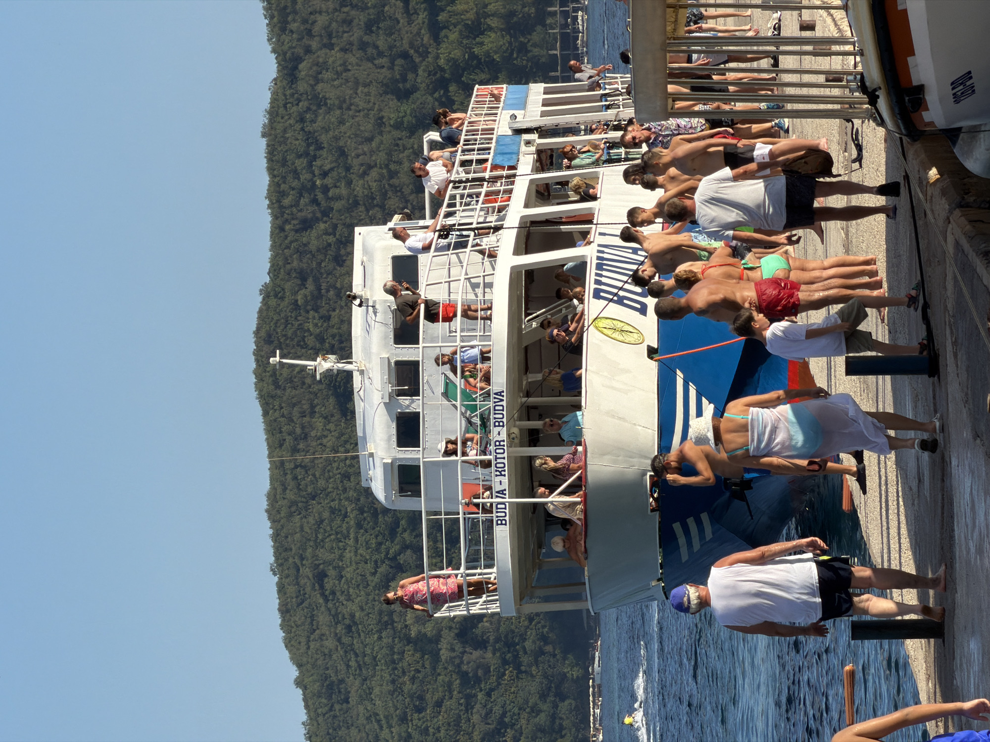 An outdoor, vertical, medium shot captures a sunny day in Budva, Montenegro, with a large passenger ferry docked at a pier. The ferry, emblazoned with "BUDVA - KOTOR - BUDVA," is the central focus, its white structure dominating the frame. Numerous people are on the ferry's decks and disembarking onto the concrete pier. Some are standing near railings, while others are seated. On the pier, a diverse group of individuals, many in summer attire, are either walking away from the ferry or standing by. The water is calm and blue, and a jet ski is visible in the distance on the left. Behind the ferry, a densely forested green hill rises under a clear blue sky. The scene suggests a tourist excursion or transport service in operation during daylight.