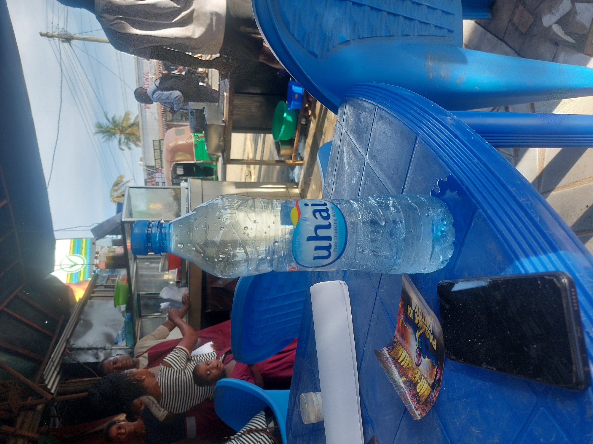 The image depicts a scene in Dar es-Salaam, Tanzania. In the foreground, a blue plastic table is prominently featured. On the table, there is a partially filled bottle of "uhai Pure Drinking Water", a rolled-up piece of white paper, a crumpled poster, and a black smartphone. Two blue plastic chairs are visible, one of which is stacked atop the other. In the background, a group of people is situated inside a modest establishment, possibly a shop or restaurant. The surroundings are urban, showing elements of the city's environment such as power lines and other buildings.