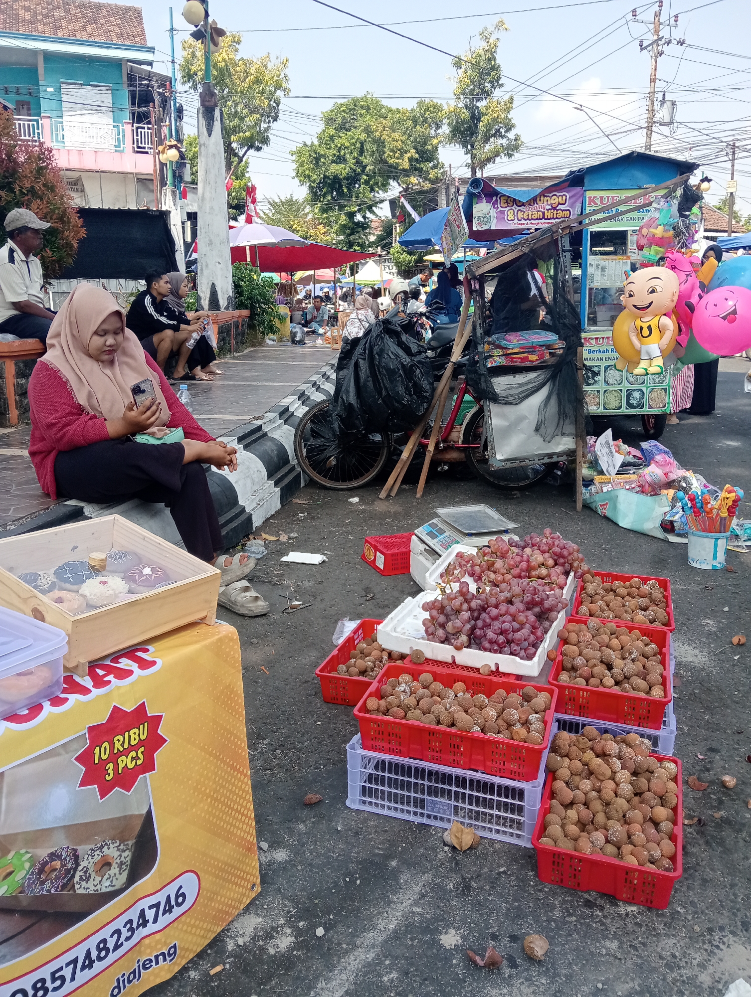 In the city of Demak, Indonesia, a busy street market unfolds under a bright sky. The scene is a vibrant mix of commerce and daily life. In the foreground, a vendor's stall displays an assortment of donuts in a wooden crate, with a prominent yellow banner announcing "10 RIBU 3 PCS" and a phone number. Next to it, red plastic crates overflow with fresh grapes and small, round fruits, likely lychees or rambutan, resting on white and grey plastic containers. A digital scale sits nearby, indicating the transactions taking place.
In the mid-ground, a woman in a hijab is seated on a low wall, engrossed in her smartphone. She wears a pink cardigan and dark pants, with sandals on her feet. Behind her, other individuals are also seated, taking a break or observing the market activity. A bicycle with a cart, laden with black plastic bags and merchandise, is parked nearby, suggesting movement and transportation of goods. A blue and white food stall, advertising "Es Dungdung & Kental Manis", stands further back, adorned with colorful balloons, including a large inflatable cartoon character. Stalls with umbrellas and various items are visible in the distance, creating a lively marketplace atmosphere. The ground is paved with asphalt, and scattered leaves suggest a natural element within the urban setting. The overall impression is one of a bustling local market, capturing the everyday commerce and social interactions of the community in Demak.