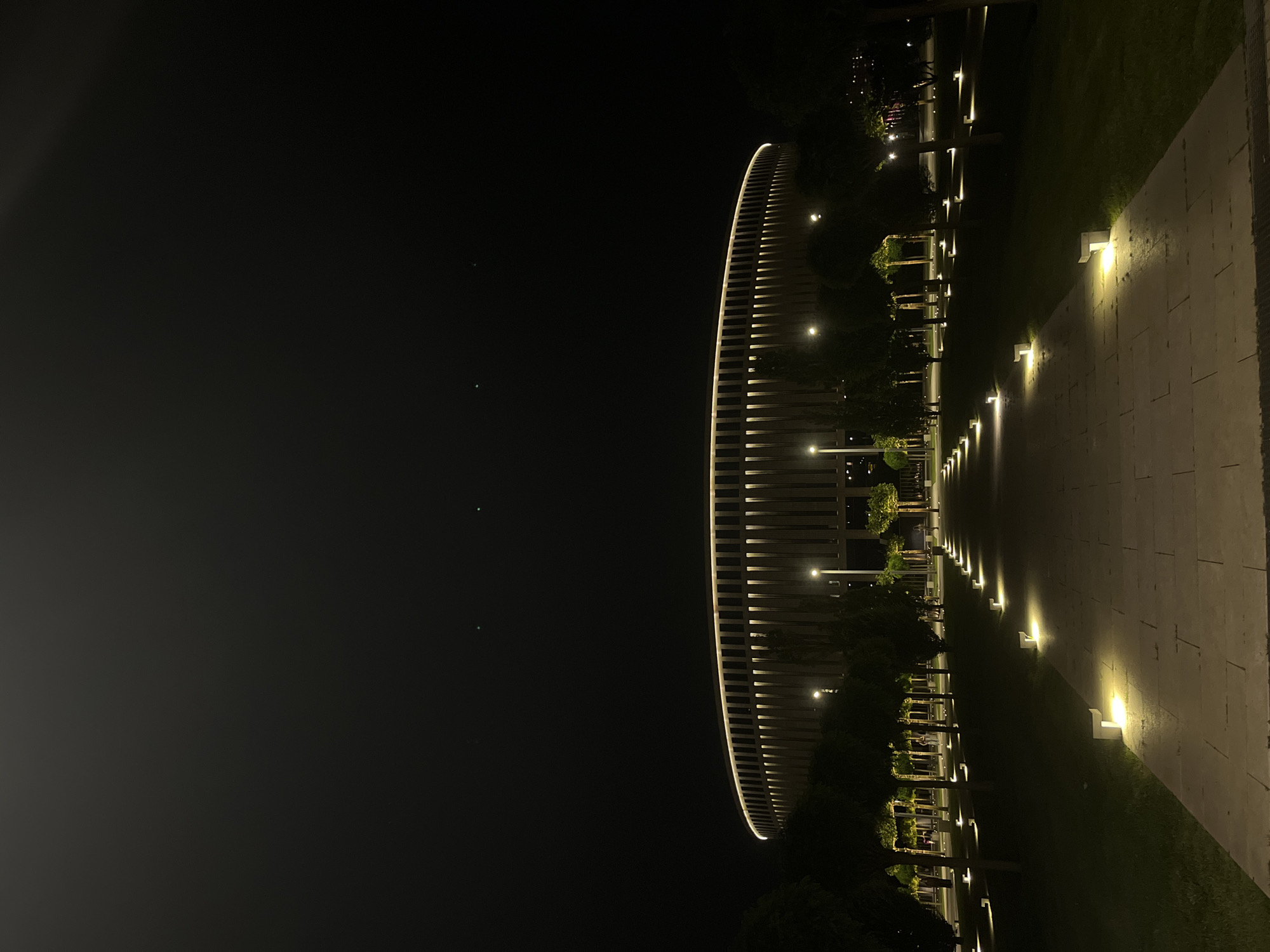 An outdoor wide shot captures a modern, circular stadium at night, illuminated by warm lighting that outlines its architectural features and creates a striking contrast against the dark sky. A pathway, lined with small, square ground lights, leads towards the stadium through a manicured lawn and rows of evenly spaced trees. The stadium's facade is characterized by vertical slats, with vertical lighting between them and a bright halo of light running along its upper edge. Two faint green lights are visible in the night sky above the stadium. The overall scene conveys a sense of serene grandeur and modern urban design, likely representing a public space in Krasnodar, Russia, possibly the FC Krasnodar Stadium.