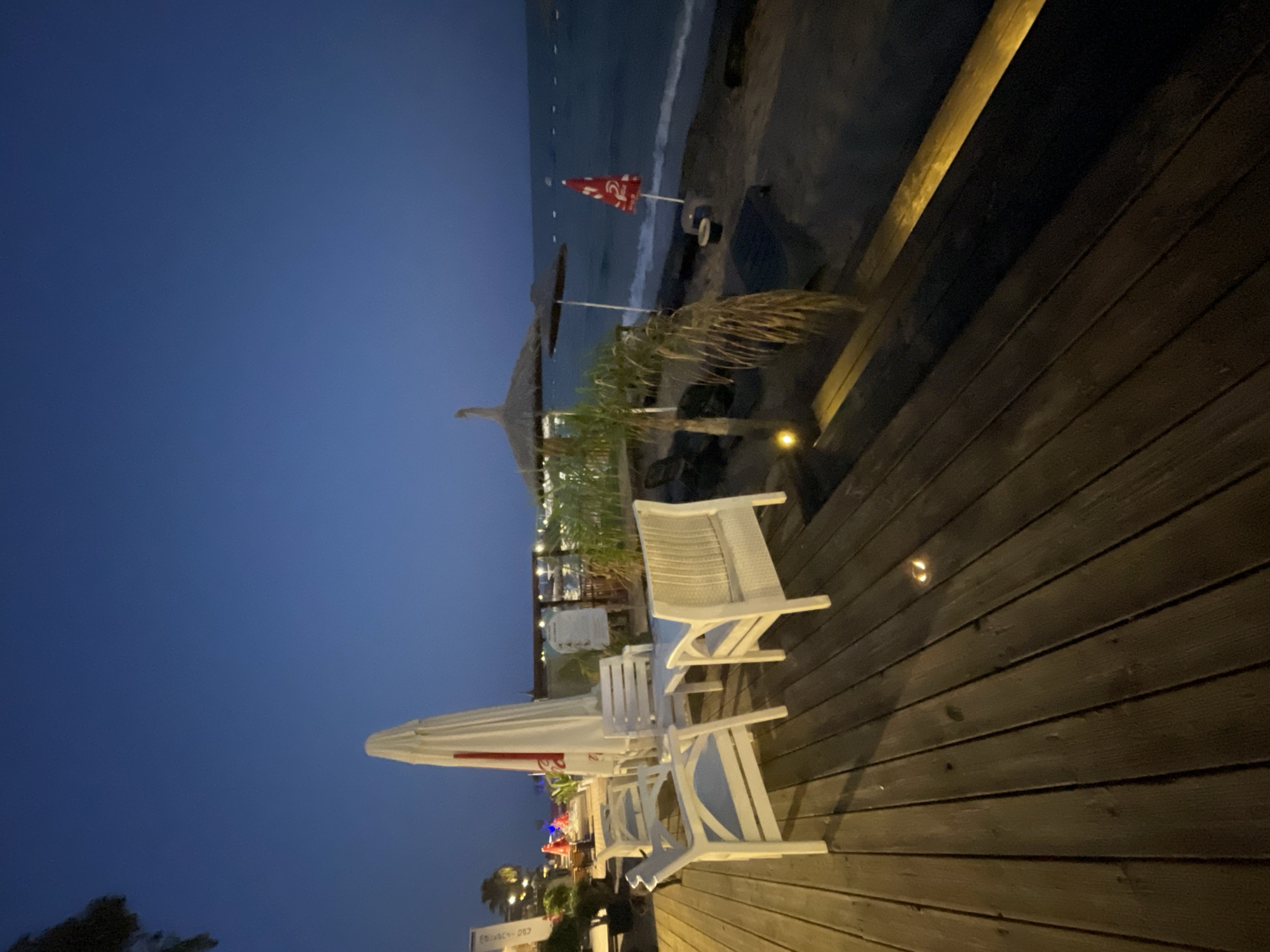 The image depicts an outdoor seating area with white furniture on a wooden deck, adjacent to a beach at dusk. The scene is located in Trikomo, Northern Cyprus. The sky is a deep blue, transitioning to a darker hue as night falls. A large white umbrella is positioned behind the seating area, and two straw umbrellas are visible on the sandy beach. The sea is calm, with gentle waves lapping the shore. The wooden deck has a warm glow from embedded lighting, and a single palm tree stands on the beach. The overall atmosphere suggests a tranquil evening at a seaside establishment.