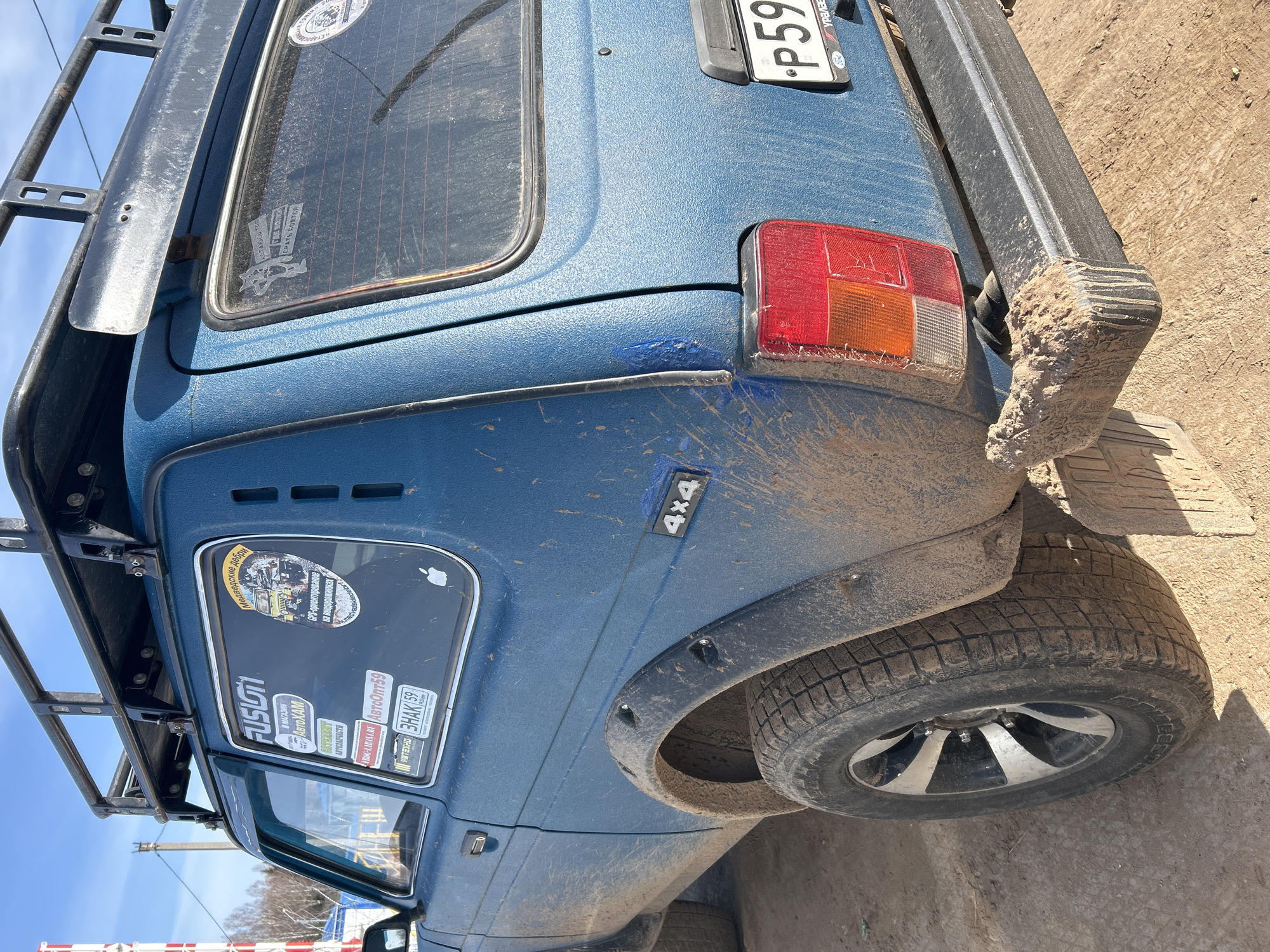 A blue Lada Niva 4x4 SUV, marked with "4x4" and splattered with mud, is partially visible, parked on a dirt surface. The rear passenger window is covered in stickers, including "FUSION", "АвтоХАМ", and "ABTOOПТ 59". There is also a sticker that reads "Медведские дебри" and a drawing with "СРАТЬ БОЯТСЯ". The license plate is partially visible and reads "P59". A metal roof rack sits on top of the vehicle. The car appears to be in Ural'skoe, Russia, as indicated by the "урала" on the license plate. The vehicle is photographed outdoors on a sunny day.