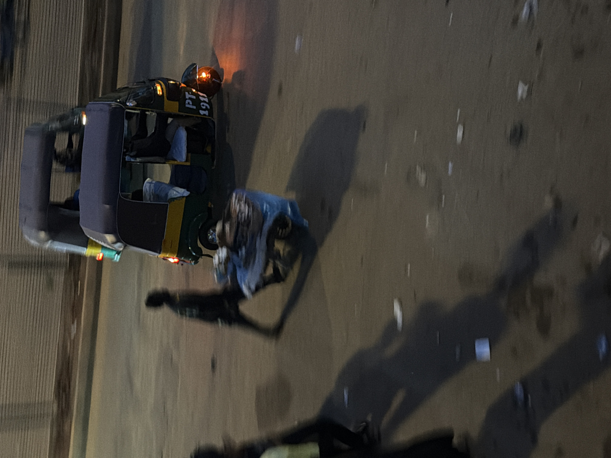 In Bukuru, Nigeria, a blurred image captures an auto-rickshaw in motion on a paved road. The vehicle, painted green and yellow, displays the license plate "PT 1911". A person is seen pushing a cart laden with goods behind the auto-rickshaw. Another individual, blurred by motion, walks in the foreground, casting a long shadow on the asphalt. The background shows a textured wall, possibly a building facade. The scene is illuminated by artificial light, suggesting it's either dusk or nighttime. Scattered white debris is visible on the road surface.