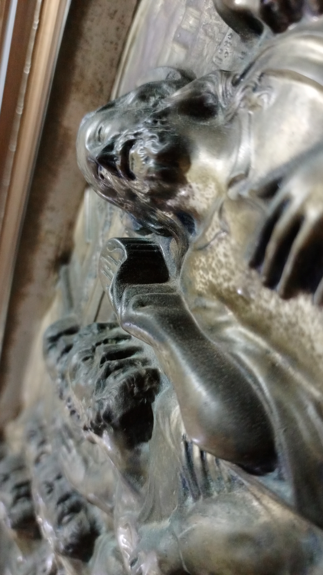 A close-up, low-angle shot focuses on a metal sculpture depicting human figures. The sculpture appears to be part of a larger piece or structure, possibly a door or panel, as suggested by the presence of a frame on the left edge. The foreground features a prominent figure with detailed facial features and hair, holding a rectangular object in their hand. The figure's expression is intense. Behind this figure are other faces, seemingly part of a crowd or procession. The metal has a dark, aged patina, with highlights that suggest a polished or reflective surface. The overall scene conveys a sense of antiquity and artistic craftsmanship. No location information regarding Los Robles, Venezuela, is discernible from the image.