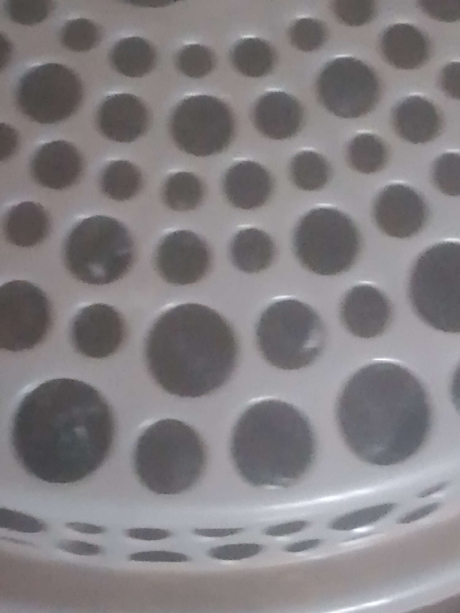 The image shows a close-up, angled view of a white plastic colander or sieve. The surface is textured with an array of raised, circular indentations or perforations of varying sizes, creating a dotted pattern. The lighting casts subtle shadows within these indentations, giving them a slightly three-dimensional appearance. The overall color palette is monochromatic, consisting of whites and grays. There are no people, animals, or any recognizable landmarks visible in the frame, and thus no specific location in Jauro Yinu, Nigeria, can be determined from the image. The focus is entirely on the abstract texture of the plastic object.