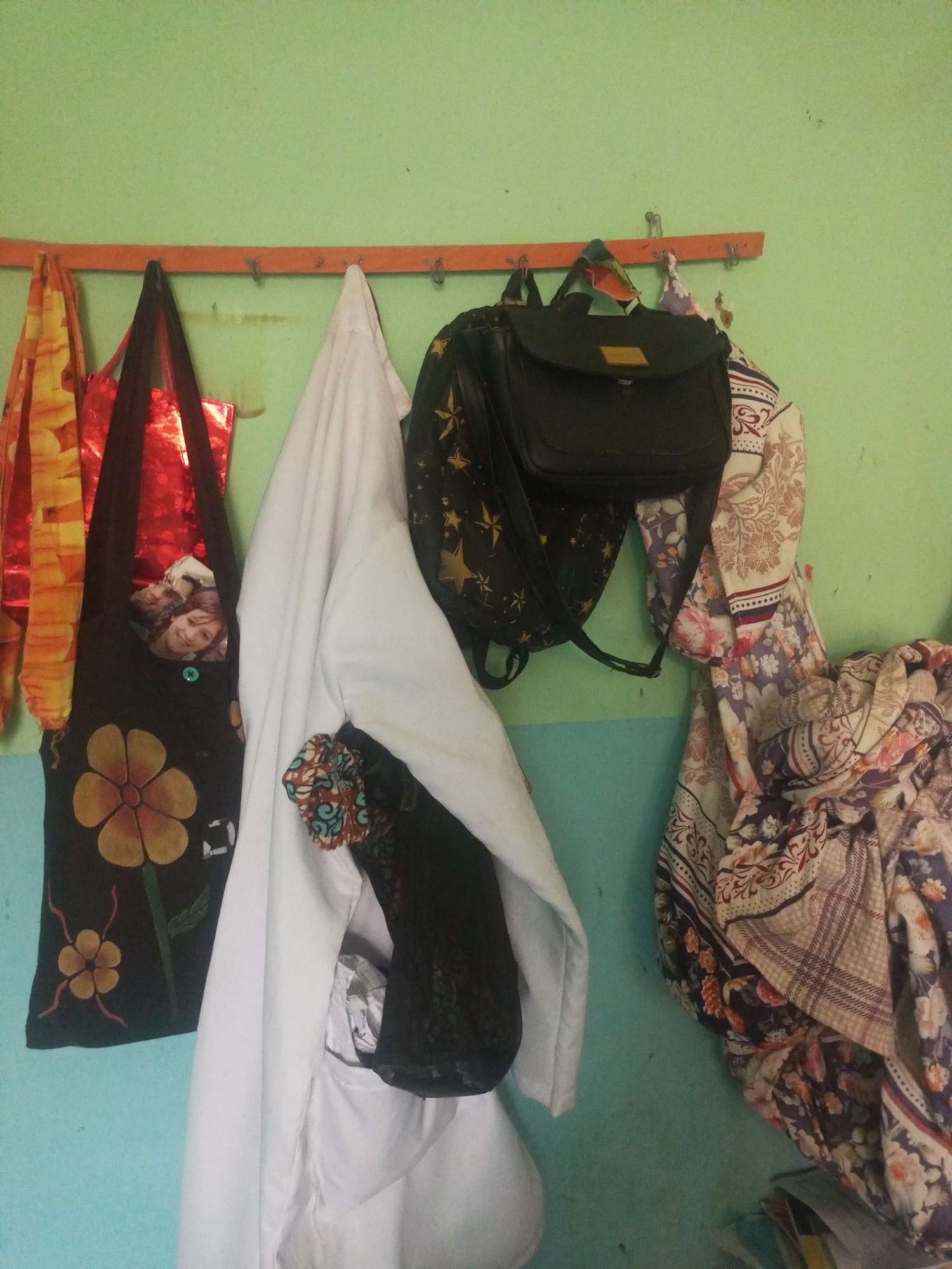 The image shows a collection of items hanging on a wall in Jalingo, Nigeria. On the left, there are two colorful fabric bags, one with a yellow and orange pattern and another red one. To the right of these, a black tote bag with a large yellow flower design and a picture of a smiling young girl is visible. Next to that, a white coat or garment is hanging, with a black patterned cloth tucked into its pocket. Further to the right, a black backpack with gold star patterns hangs alongside a large, elaborately patterned textile. The items are suspended from a horizontal orange rack mounted on a pale green wall. The lower portion of the wall transitions to a light blue color. The scene appears to be indoors, likely in a domestic or storage space.