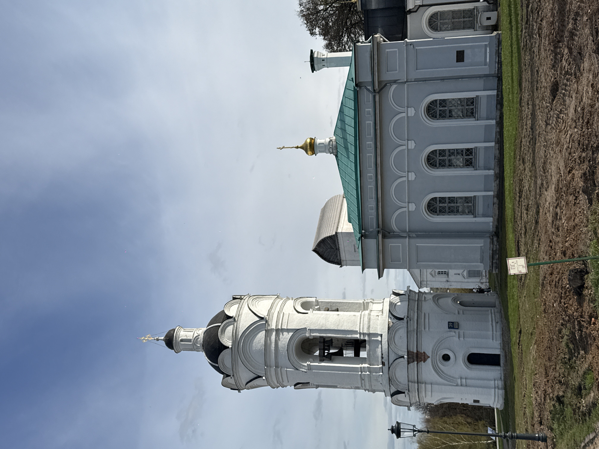 The image depicts the Church of the Ascension in Kolomenskoye, Moscow, Russia. The church is constructed of white stone and has a unique tent-like roof structure. It is located in a park-like setting with grass and trees surrounding it. A grey building is visible to the left, and a pole with a sign is visible to the right. The sky is cloudy.