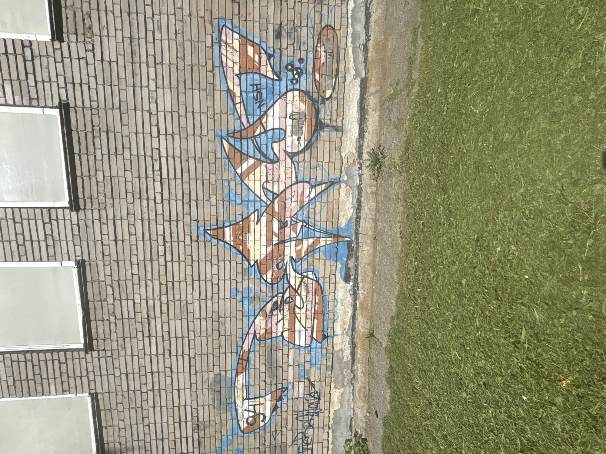 An exterior shot captures a section of a light gray brick wall with two windows in the upper portion. Below the windows, graffiti in blue, orange, and white colors adorns the wall. The graffiti features stylized lettering and abstract shapes. In the lower half of the image, a strip of cut green grass extends horizontally across the scene, with a concrete walkway visible between the grass and the wall. The background consists of the brick wall. The image appears to be taken in an urban outdoor environment.