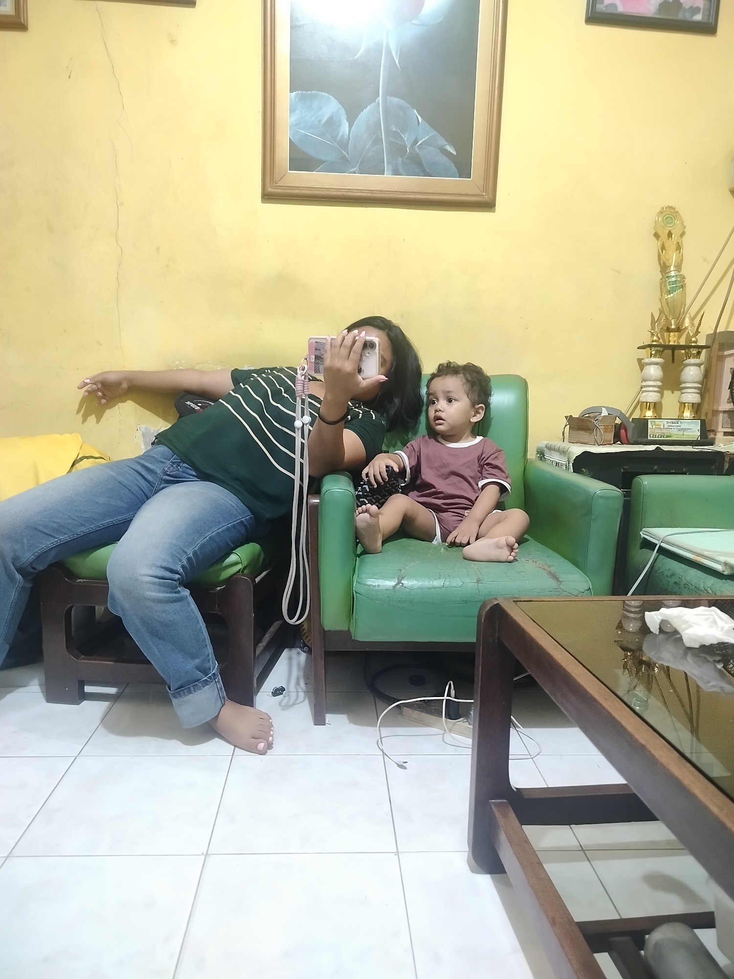 In a domestic setting in Surabaya, Indonesia, a young child with curly brown hair, wearing a maroon t-shirt and shorts, sits on a green sofa. To their left, an adult, partially obscured, is lying down, holding a smartphone and appearing to take a selfie. The adult is wearing a dark green t-shirt with white stripes and blue jeans. The room is characterized by a yellow-painted wall with visible cracks, a framed picture of a rose on the wall, and a trophy stand. A wooden coffee table with a glass top is positioned in the foreground. The floor is tiled with white square tiles.