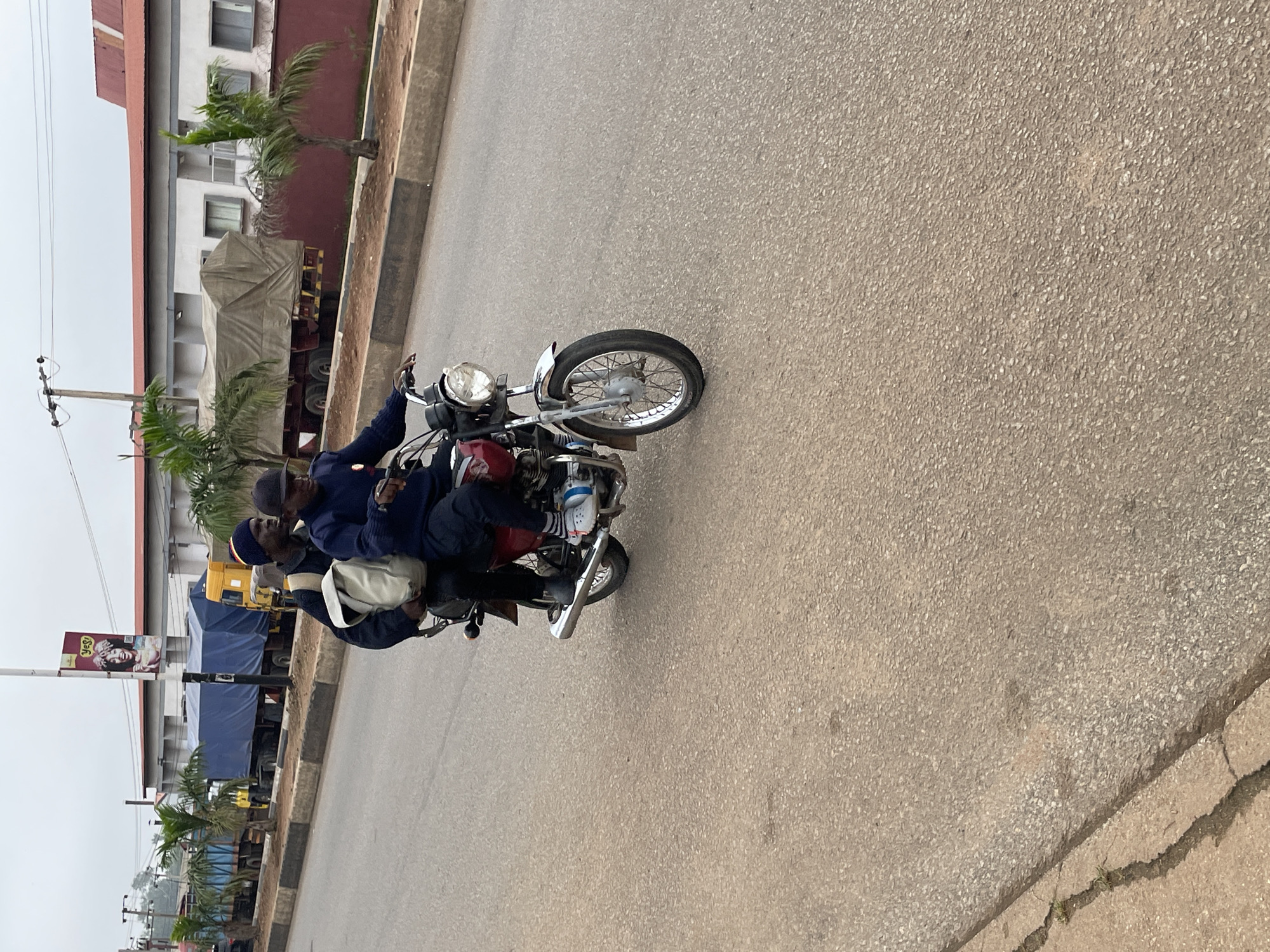 Two individuals are riding on a red motorcycle on a paved road. The person in the front, wearing a dark blue long-sleeved shirt and a black cap, is operating the motorcycle. The person in the back, wearing a dark blue long-sleeved shirt and a blue and red beanie, has a light-colored bag slung over their shoulder. The road has a rough, asphalt surface. In the background, there is a row of parked trucks with blue tarps, a yellow truck, and buildings. Palm trees are visible between the trucks and the buildings. A billboard with an image of a person is attached to a pole behind the trucks. The sky is overcast and appears to be daytime. The scene is located on a street in Barakin Lerawi, Nigeria.