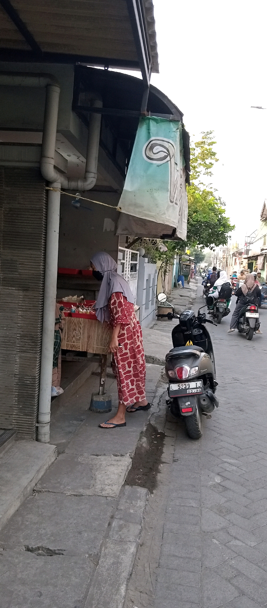 A person wearing a gray hijab and a red and white patterned dress is standing in front of a wooden stand displaying various items, possibly food or goods, in Wadungasri, Indonesia. The individual appears to be looking at or arranging items on the stand. The scene is on a street with concrete pavement and a gutter. Several motorcycles are parked or being ridden in the background, indicating a public street setting. A green banner with a black and white swirl design hangs from an overhead structure. The surrounding buildings are simple structures, and trees are visible in the background. The lighting suggests it is daytime.