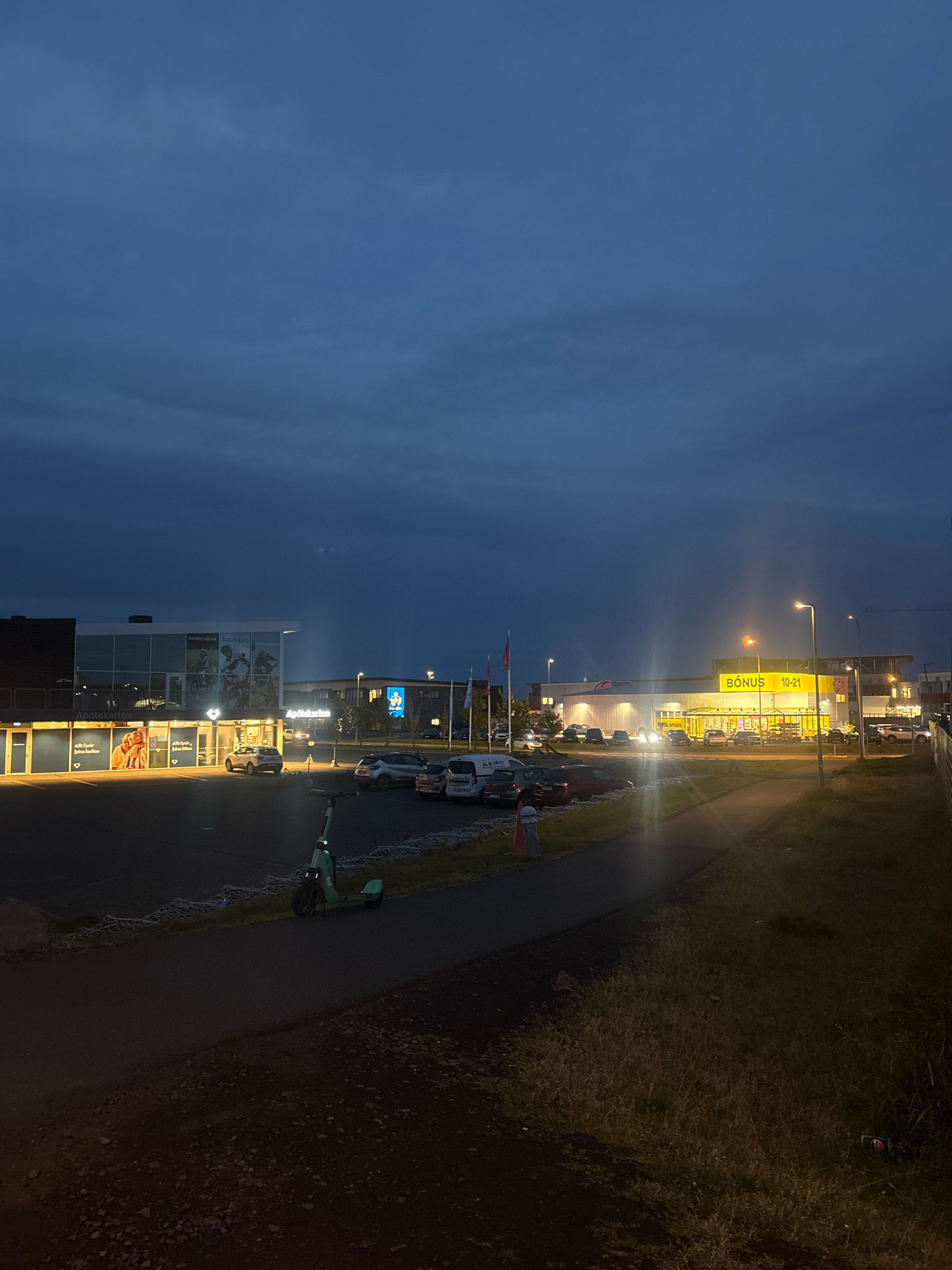 The image depicts a street scene in Reykjavik, Iceland, during twilight. The sky is a deep, dusky blue with visible clouds. On the left, a modern building complex features businesses with illuminated signs, including "Apótekarinn" (pharmacy) and another with images of people cycling. A row of parked cars is visible in front of this building.
To the right, a brightly lit "BÓNUS" supermarket stands out with its distinctive yellow signage, indicating opening hours of "10-21." Several cars are parked in the supermarket's lot, and streetlights illuminate the area.
In the foreground, a paved pathway runs along the edge of a grassy area. A green electric scooter is parked on the pathway. The overall impression is a commercial area of Reykjavik at the end of the day.