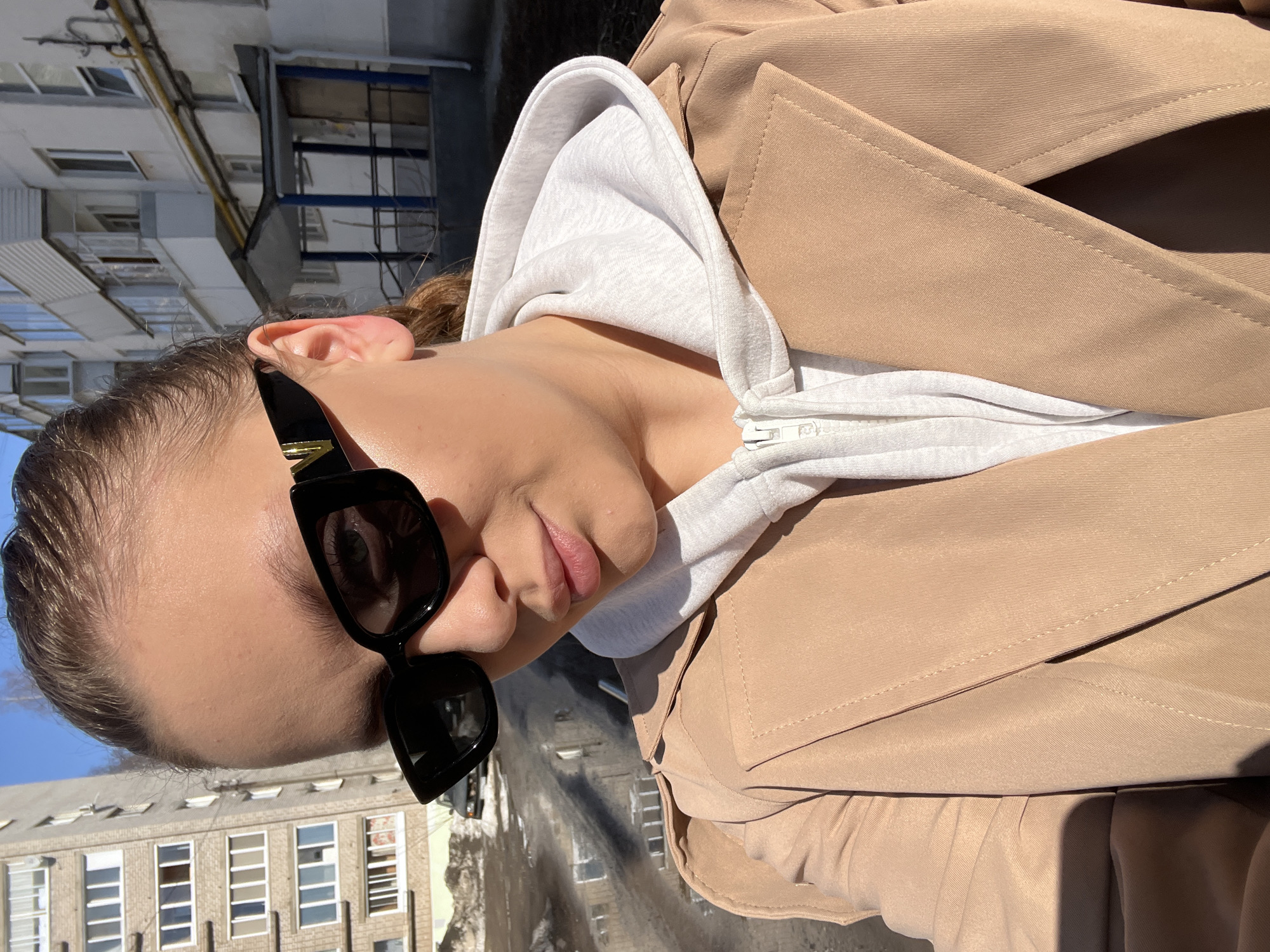A woman wearing sunglasses, a light gray hooded sweatshirt, and a tan trench coat is the primary subject of the image. She is positioned in front of a background consisting of multi-story buildings. Based on the architecture, specifically the design of the buildings in the background, this location is potentially in the city of Chaikovsky, Russia.
