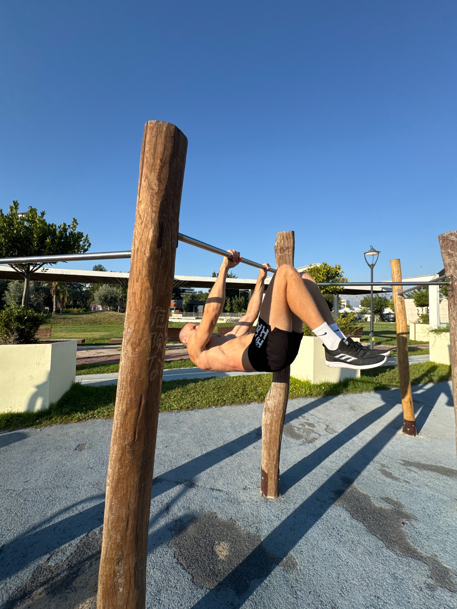 The image captures a man performing a hanging leg raise exercise on an outdoor fitness equipment in Antalya, Turkey. The man is shirtless and wearing black athletic shorts and white socks with black sneakers. He is suspended from a metal bar supported by two wooden posts. The background reveals a park-like setting with lush green trees, paved walkways, and some modern architecture. The sky is clear and blue, indicating a sunny day. The lighting suggests it's either early morning or late afternoon due to the long shadows cast by the wooden posts on the blue-colored ground. The fitness equipment appears to be made of natural wood and metal, designed for outdoor use in a public space.
