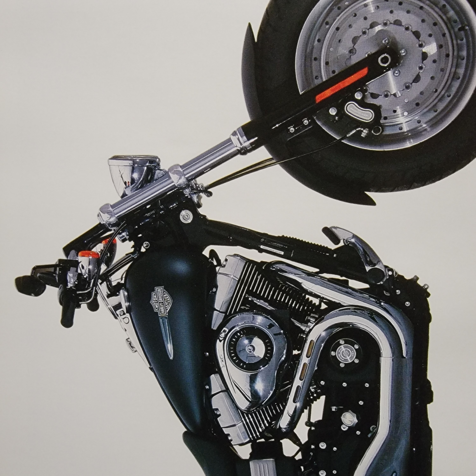 The image is a close-up, right-side, three-quarter view of a black Harley-Davidson motorcycle parked against a plain white background. The motorcycle is the sole subject, with its front wheel, fender, front suspension, engine, fuel tank, and handlebars visible. The engine appears to be a V-twin configuration with prominent chrome accents, including the air intake cover and exhaust pipes. The fuel tank has a Harley-Davidson logo. The front wheel is a disc brake with a chrome rim and black tire. The motorcycle is positioned slightly to the left of the frame, with the front wheel angled slightly towards the viewer. The lighting is even and bright, suggesting a studio setting. There are no people or any other objects in the frame, and the location is specified as Unknown, Unknown.