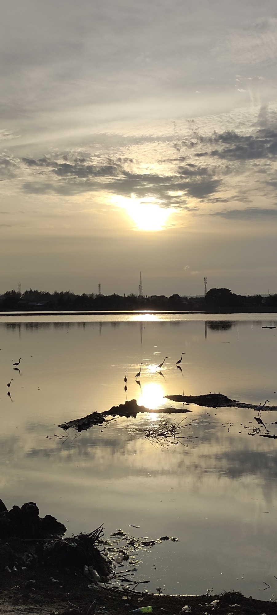 This is a vertical, wide-angle, outdoor, daylight photograph taken at the Lhokseumawe Reservoir in Lhokseumawe, Indonesia. The sun is visible in the upper central portion of the frame, partially obscured by clouds, casting a warm, golden light across the scene. The reservoir's surface is calm and reflective, mirroring the sky and the silhouettes of several birds wading in the shallow water. The birds appear to be egrets or herons, with long legs and necks. In the background, a treeline separates the water from a hazy horizon featuring industrial structures, likely communication towers, suggesting the presence of urban development. The foreground shows a murky, debris-strewn shoreline. The overall impression is one of a tranquil natural setting juxtaposed with industrial elements.