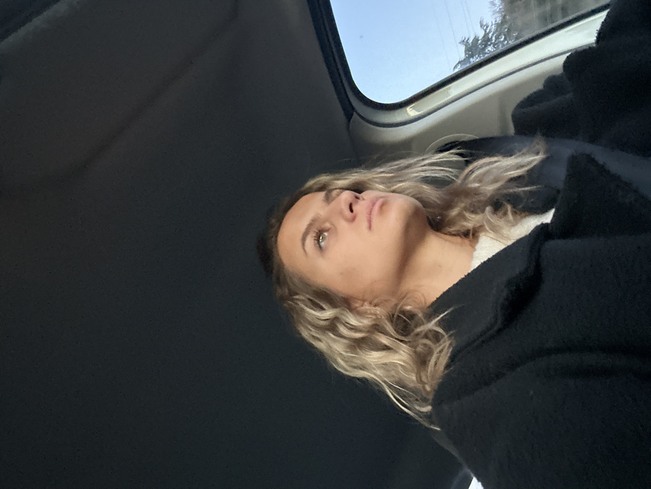 A young woman with wavy blonde hair and fair skin is looking upward, presumably out of the sunroof of a car. She is wearing a black coat over a white top. The view outside the sunroof shows a blue sky and a glimpse of trees. The interior of the car is mostly dark, with the roof dominating the left side of the frame.