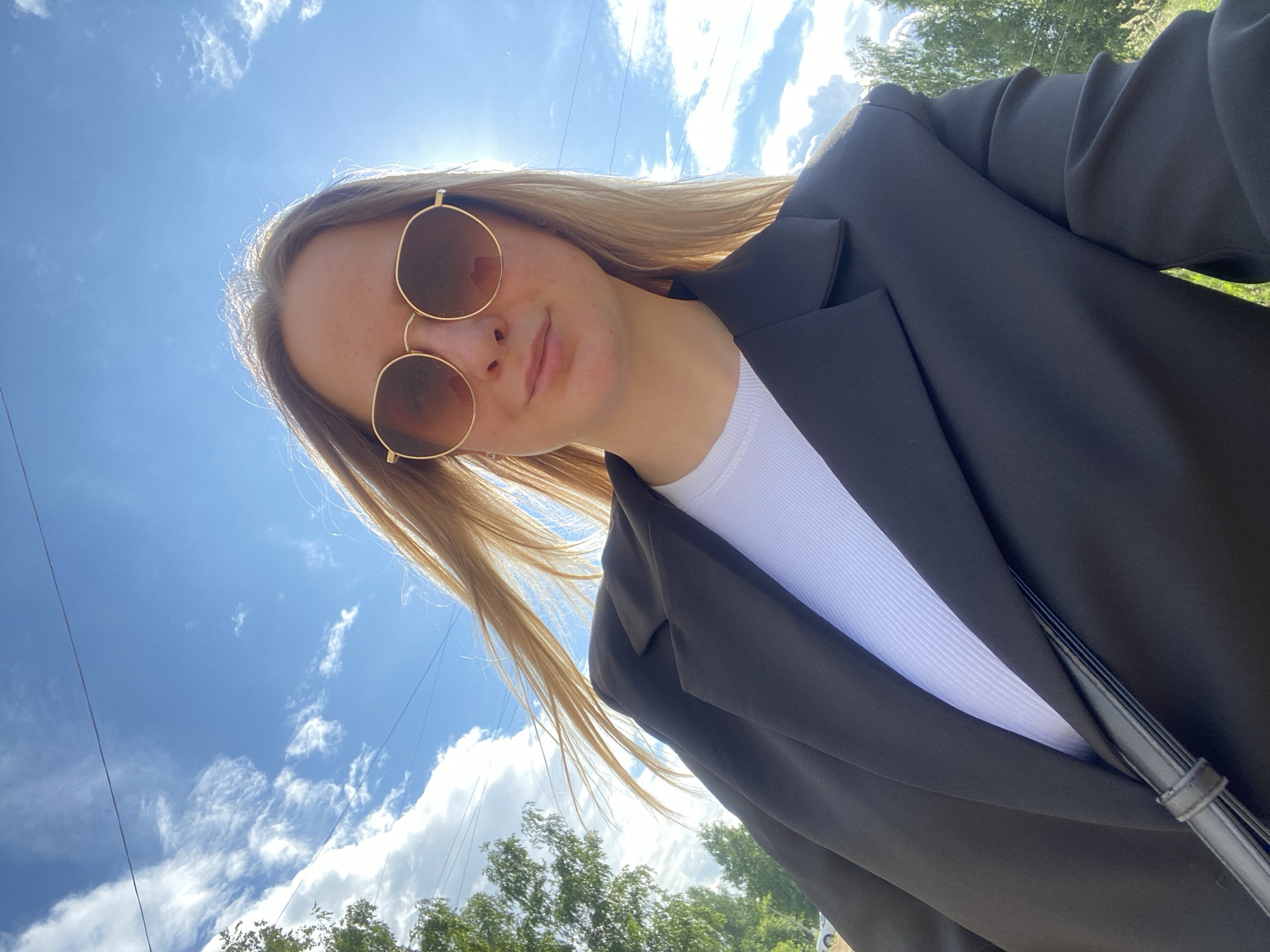 A young woman with blonde hair and sunglasses is taking a selfie outdoors on a sunny day. She is wearing a black blazer over a white ribbed top. Her hair is blowing in the wind. In the background, there is a bright blue sky with scattered white clouds, and some greenery, possibly trees. The image was taken in the city of Chaikovsky, Russia.