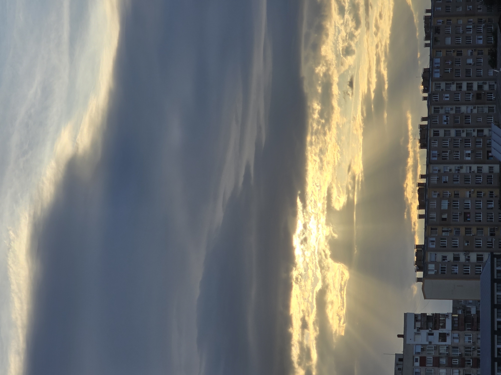 The image depicts a sunset over a cityscape, likely taken from a high vantage point. The sky is filled with clouds, some illuminated by the setting sun, creating a golden glow. To the right, high-rise residential buildings with numerous windows form a dense urban landscape. The perspective suggests the location is in a city, though specific details about the city are not provided.