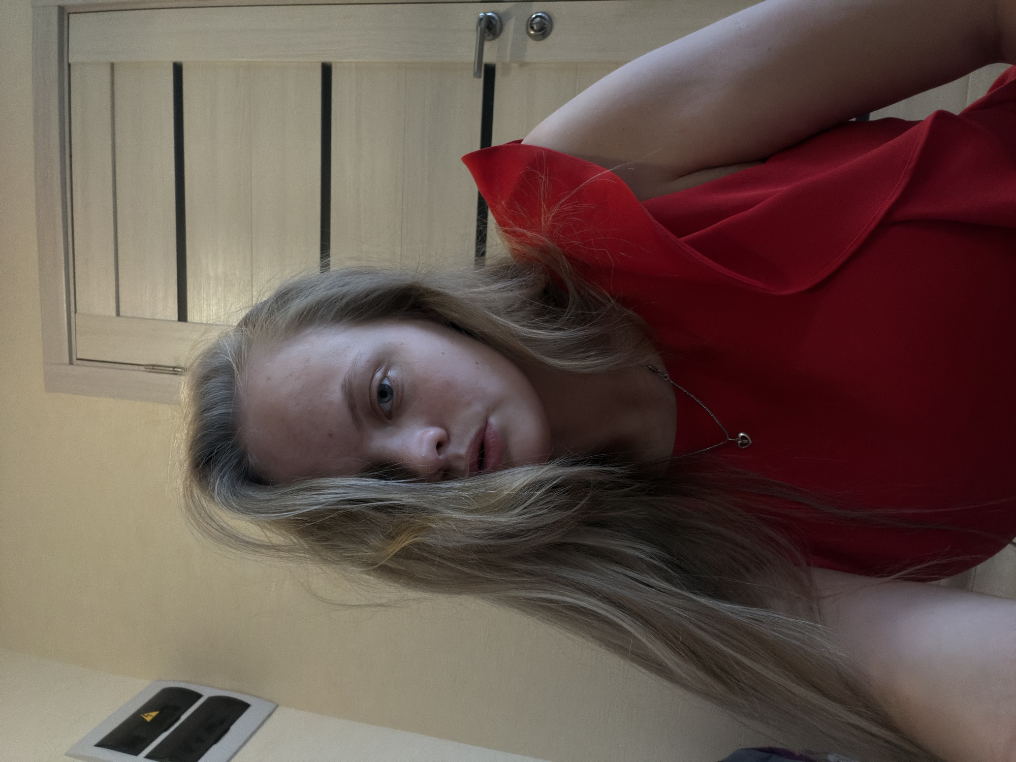 A close-up portrait shows a young woman with long, straight blonde or light brown hair looking directly at the camera. The image is rotated 90 degrees counter-clockwise. She has light blue or grey eyes and a fair complexion, with some blemishes visible on her face. She wears a bright red top or dress featuring a ruffled neckline, and a delicate silver-colored necklace with a small heart pendant. Her left arm is visible, bent and positioned behind her head or shoulder.
The background depicts an indoor setting, including a light-colored wooden door with vertical paneling and two silver doorknobs or handles. Below her face, on a flat surface, there is a small white item with two black rectangular objects, one of which displays a yellow triangular symbol containing a black lightning bolt, indicating an electrical warning or hazard. The lighting appears soft, suggesting an indoor shot during the day. The woman's expression is neutral to slightly contemplative. The scene conveys a personal moment, likely a selfie, in a domestic environment in Izhevsk, Russia.