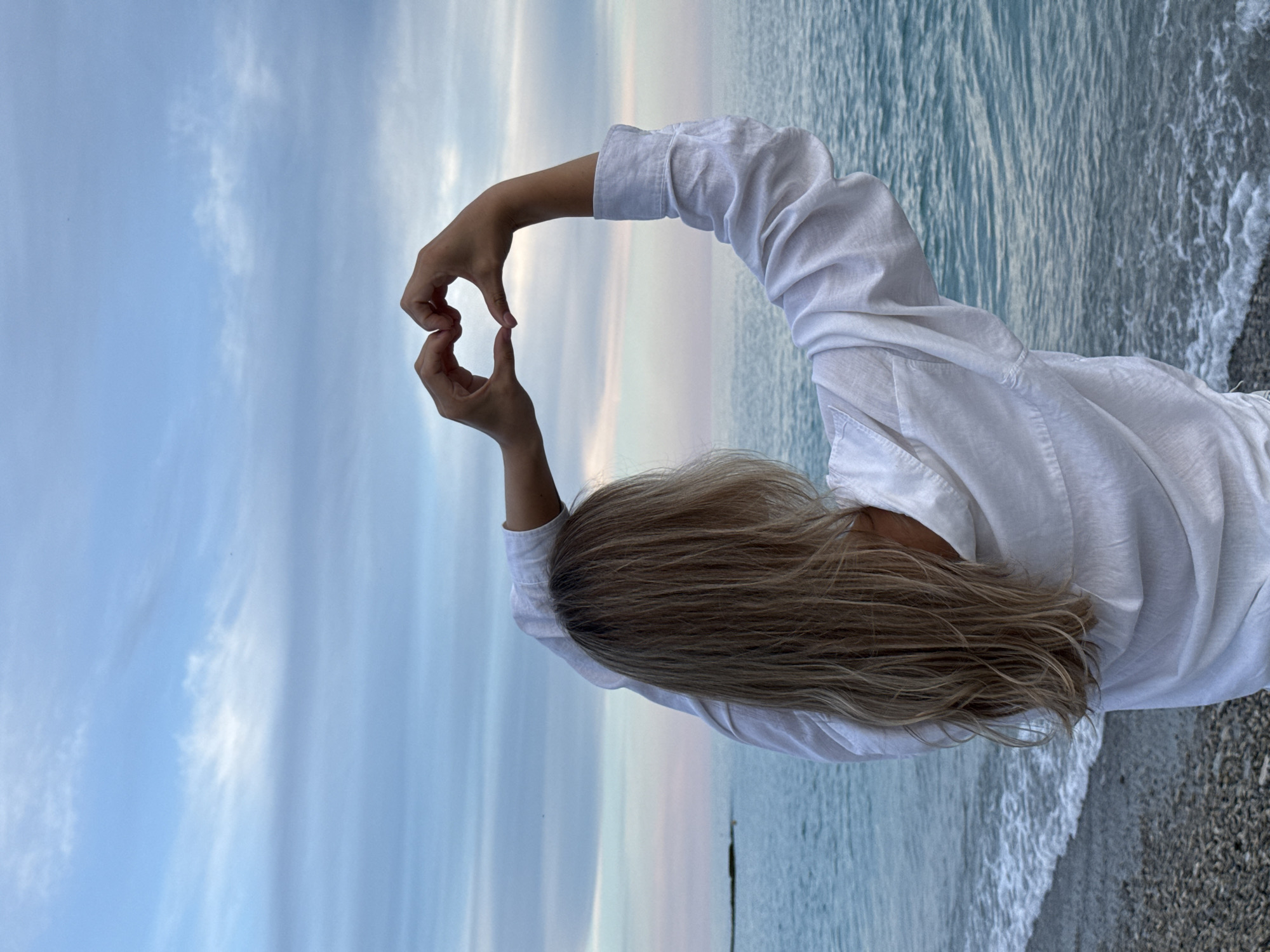 A person with long blonde hair, seen from behind, stands on a pebbly beach facing the sea, making a heart shape with their hands. The person is wearing a white long-sleeved shirt. The sea is calm with small waves lapping at the shore. The sky is filled with soft, pastel-colored clouds. This scene is located on the coast of the Black Sea in the village of Nebug, Russia.